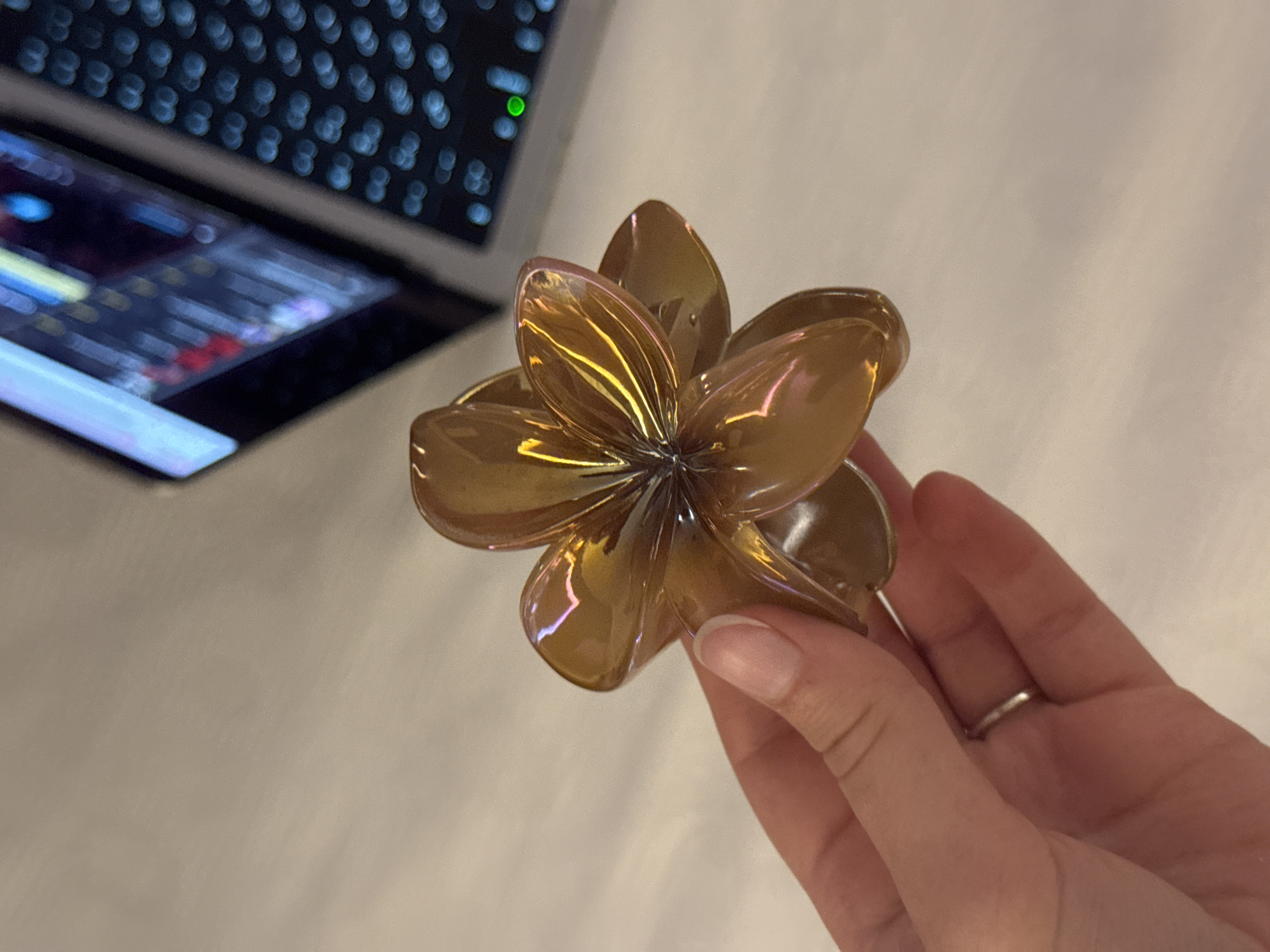 A close-up, high-angle shot shows a person's left hand holding a translucent, amber-colored, five-petaled flower-shaped ornament. The ornament has a glossy finish and reflects light, showing iridescent pink and yellow hues. The hand has clean fingernails and a thin silver ring on the ring finger. In the background, a blurred laptop is visible with its screen displaying what appears to be code or data with various colored segments. The surface on which the laptop rests is light-colored and textured, resembling a desk. The scene is indoors, likely in an office or home environment, and there is no indication of the city of Aksu, Turkey, within the visual information provided.