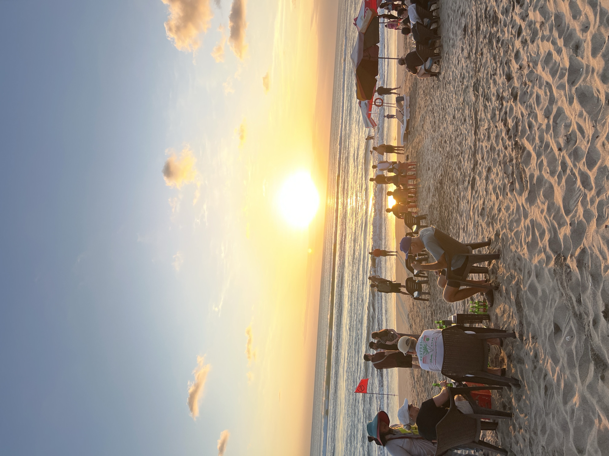 This is a photograph taken at sunset on Kuta Beach, Bali, Indonesia. The sun is setting over the ocean, casting a warm glow across the sky and the sand. A group of people are sitting at tables on the beach, enjoying drinks and conversation. Some are facing the ocean, while others are looking at each other. A red flag is planted in the sand near the water's edge. There are also beach umbrellas and lounge chairs visible in the background. The waves are gently rolling in, and the overall atmosphere appears relaxed and festive. The sand is a light golden color, and it is covered in footprints and small dunes. The sky is a gradient of orange, yellow, and blue, with a few scattered clouds. The ocean is a deep blue, with white caps on the waves. The scene is typical of a beach sunset experience in Kuta, a popular tourist destination known for its vibrant nightlife and beautiful beaches.