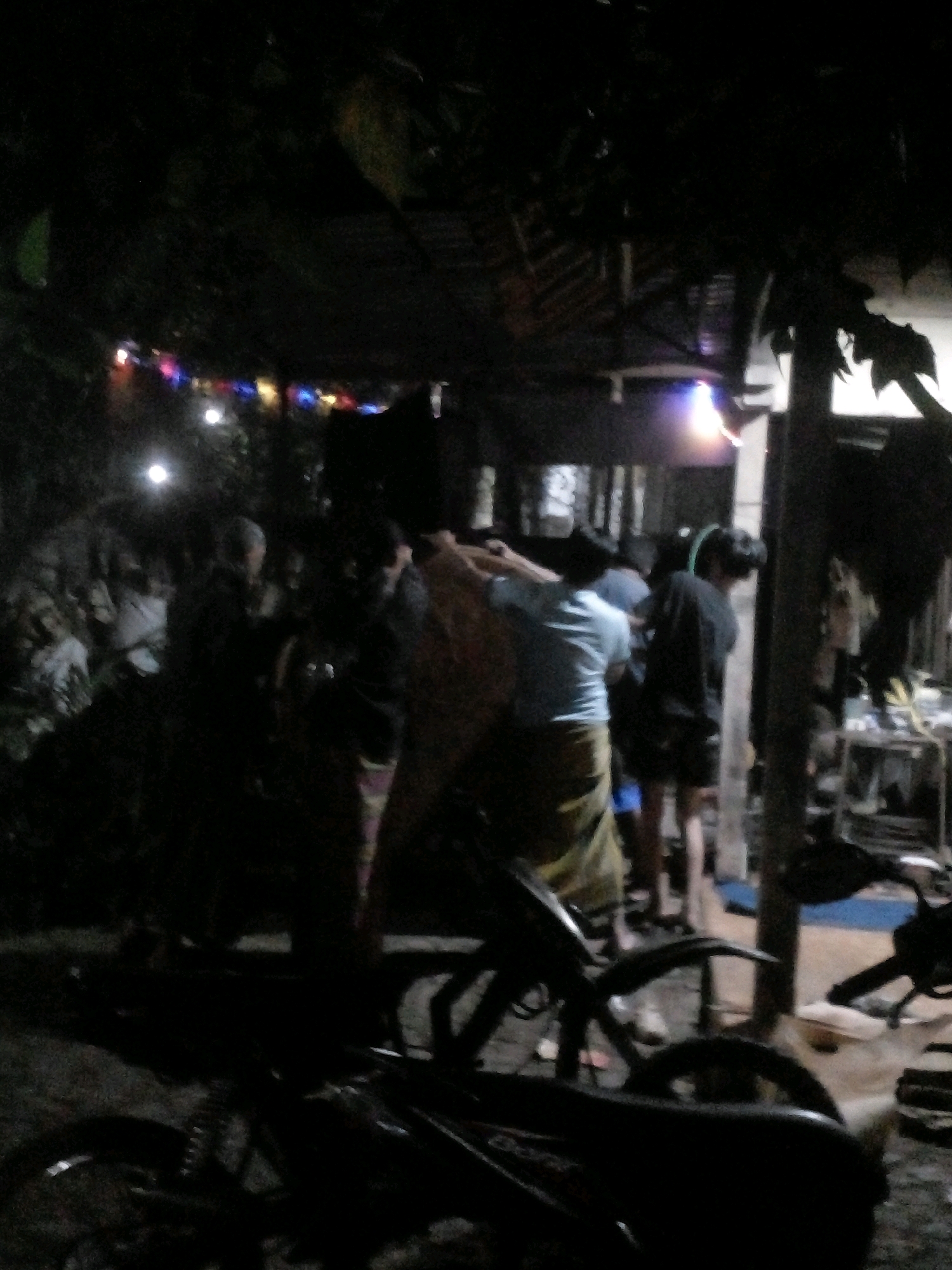 The image depicts a nighttime scene in Pungangan, Indonesia, appearing to be a gathering in a residential or community area. Several individuals are present, many obscured by darkness. In the foreground, a motorcycle is parked. Behind the motorcycle, a group of people are clustered, some appearing to be engaged in conversation or activity. Decorations, possibly string lights or banners, are visible in the background, suggesting a festive occasion. The overall environment suggests an outdoor setting, likely a yard or street, with some structures visible in the distance. The lighting is dim, with artificial lights contributing to the illumination.