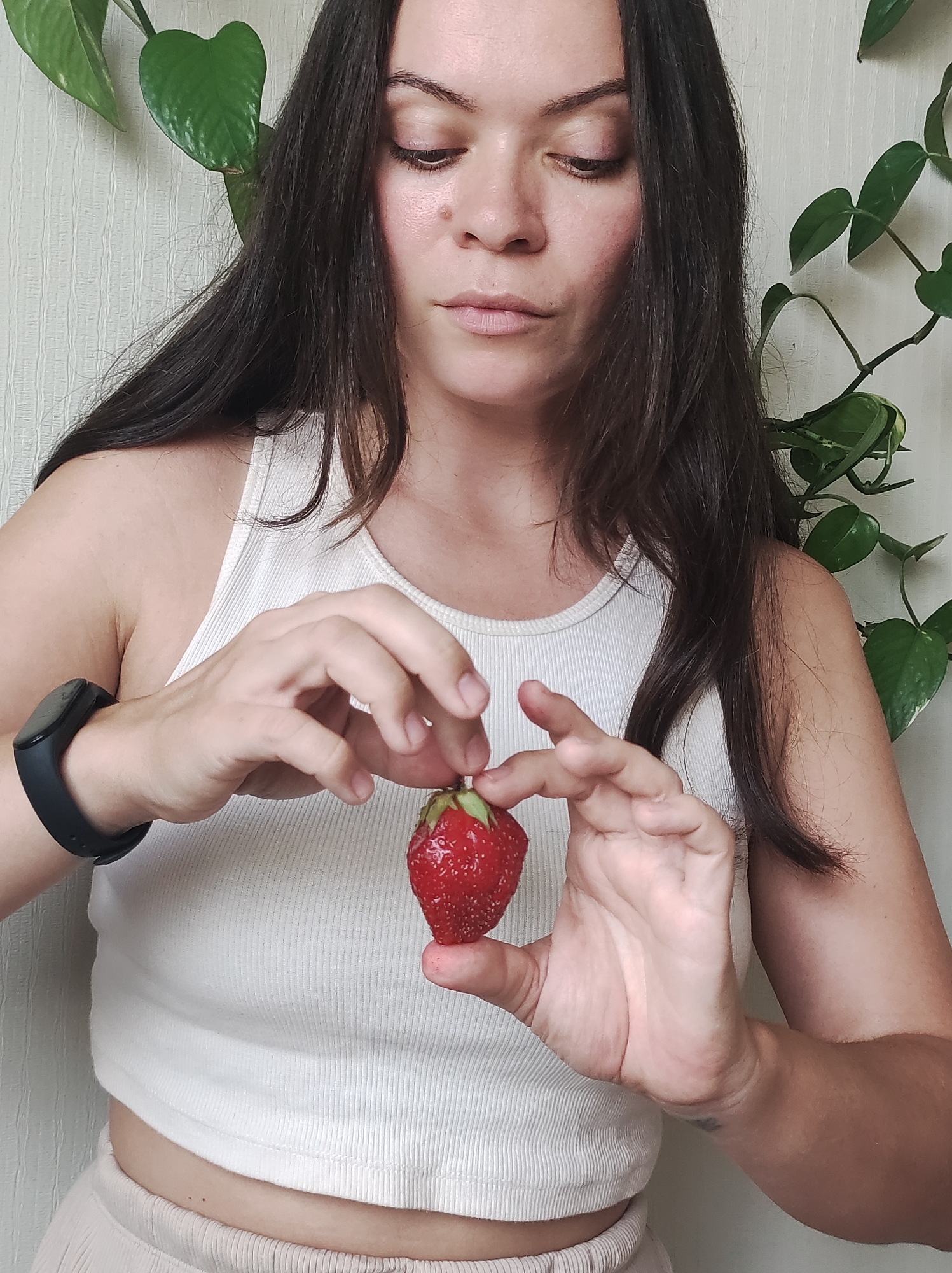 A woman with long, dark hair wears a white tank top and light-colored pants. She is holding a single red strawberry between her fingers and thumb, examining it. She has a dark-colored fitness tracker on her left wrist. Behind her, a green plant with large leaves grows against a light-colored, textured wall. The setting appears to be indoors, possibly a residential room in Chaikovsky, Russia.