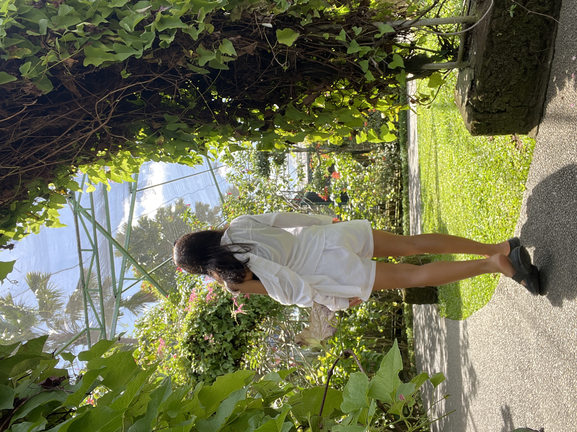 A woman in a white dress stands in a garden in Sukawati, Indonesia. She faces away from the camera. The garden has lush greenery and a structure with vines. The path she stands on is paved.