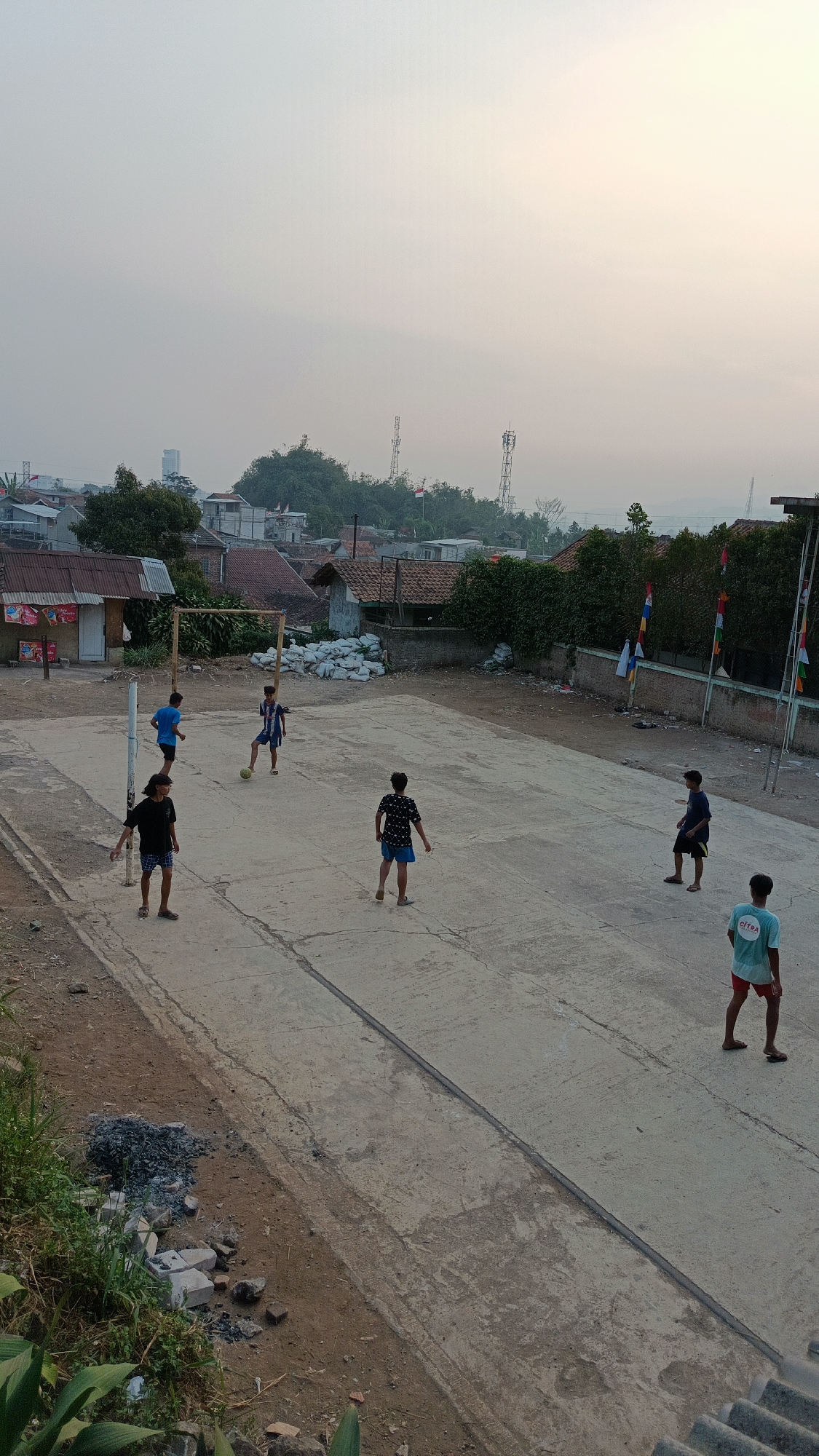The image depicts an outdoor concrete court, possibly a sports field, in Margajaya, Indonesia. Six young males are present on the court, engaged in a game of soccer. Two individuals are in the background, one in a blue shirt and shorts, the other in a blue and white striped shirt and blue shorts. In the foreground, four individuals are active: one in a black shirt and shorts to the left, another in a patterned black and white shirt and blue shorts in the center, a third in a blue shirt and dark shorts to the right, and a fourth in a teal shirt with "CITRA" printed on it and red shorts, also on the right. A soccer goal constructed of wooden poles is visible on the left side of the court. Surrounding the court are buildings with corrugated metal roofs, a brick wall adorned with colorful flags, and scattered piles of white sacks. The background reveals a densely populated residential area with various structures, hills covered in greenery, and telecommunication towers. The sky is overcast, suggesting it may be early morning or late afternoon. The overall impression is a casual, community sporting activity in a somewhat undeveloped urban or semi-urban setting.