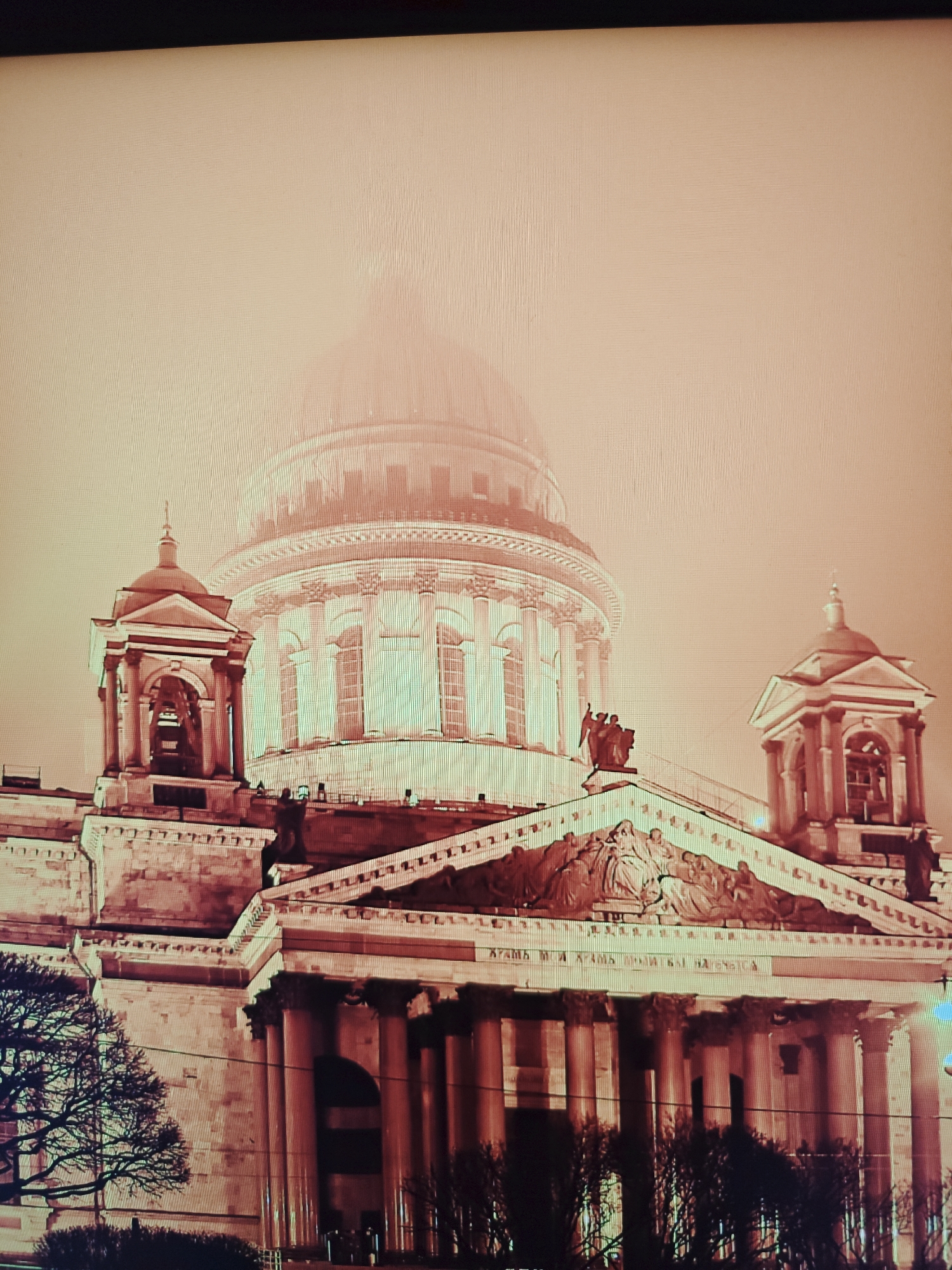The image shows a large, ornate building with a central dome and two smaller domes on either side. The building is made of stone and features numerous columns and architectural details. A sculpture of figures is visible on the facade. Trees are present in the foreground. The overall tone of the image is reddish, suggesting it may be a nighttime or sepia-toned photograph.