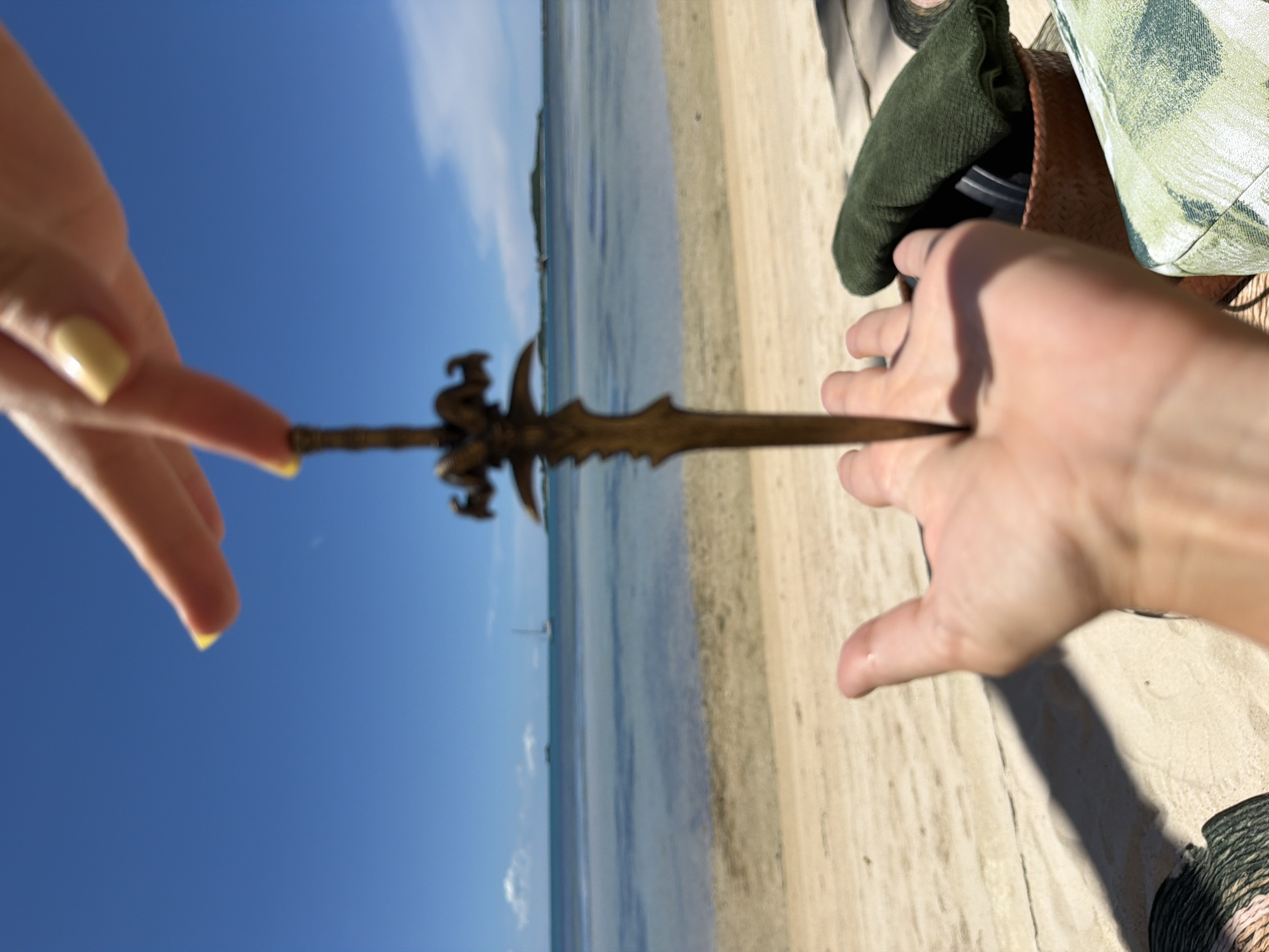 In Baan Thong Krut, Thailand, a person's hands are holding a miniature sword over a sandy beach. The sword is brass-colored and has a decorative hilt with ram-like horns. The right hand, with yellow-painted fingernails, is positioned above the sword, with two fingers extended. The left hand, palm up, is positioned below the sword, appearing to catch it. The background shows a calm, blue ocean with two sailboats in the distance, and a clear, bright blue sky. Part of a woven basket containing a green cloth and other objects, and a patterned fabric item, are visible on the right side of the frame, suggesting the scene is set near a lounging area.