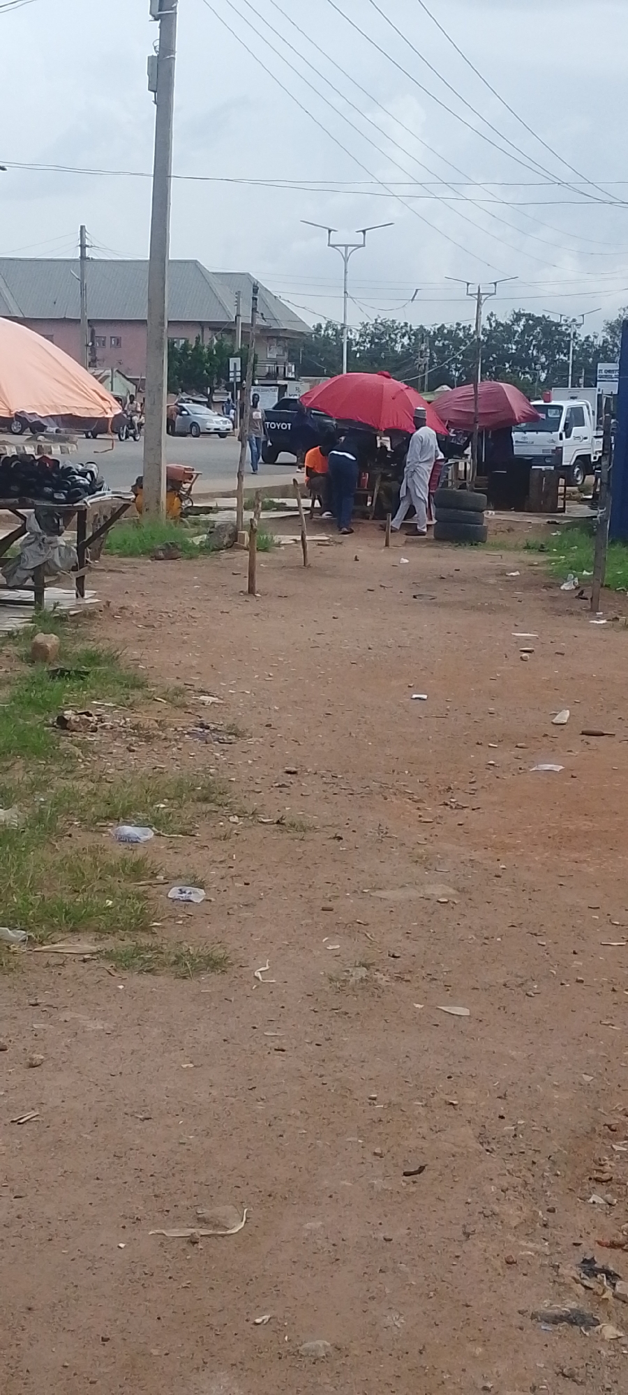 The image captures a street scene in Gura Top, Nigeria, under an overcast sky, suggesting it might be daytime with cloudy weather. The foreground is a dirt path littered with various debris, including plastic bottles and what appears to be discarded paper. In the middle ground, a bustling market scene unfolds. Several people are visible, some engaged in conversations or tending to their wares.
A prominent feature is a stall with an orange canopy, displaying a collection of black shoes. Nearby, a table also holds shoes, implying a shoe vendor. To the right, a makeshift setup with red and maroon umbrellas provides shade for another stall. Underneath these umbrellas, people are gathered, possibly customers and vendors. One individual, dressed in a white outfit and a cap, walks past. Another person in an orange shirt is seated, and someone in dark clothing stands nearby. Stacked tires are visible next to this stall, hinting at a possible tire repair or sale business.
In the background, vehicles are present. A silver car is seen on the road, and a black Toyota pickup truck is parked. Beyond the market area, buildings with tiled roofs and windows are visible, suggesting a developed settlement. Streetlights with solar panels are mounted on poles, indicating a modern infrastructure element within the otherwise rustic setting. The overall impression is one of a lively local market, a common sight in many Nigerian towns. There is no discernible text in the image apart from the "TOYOT" logo on the pickup truck.