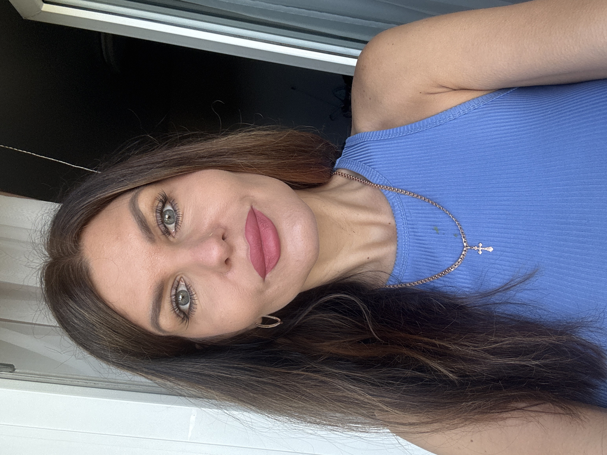 A woman with brown hair and green eyes is wearing a blue ribbed tank top and a gold chain with a cross pendant. She is wearing lipstick and earrings. She appears to be standing in front of a window. There is insufficient information to determine the location in the city of Chaikovsky, Russia.