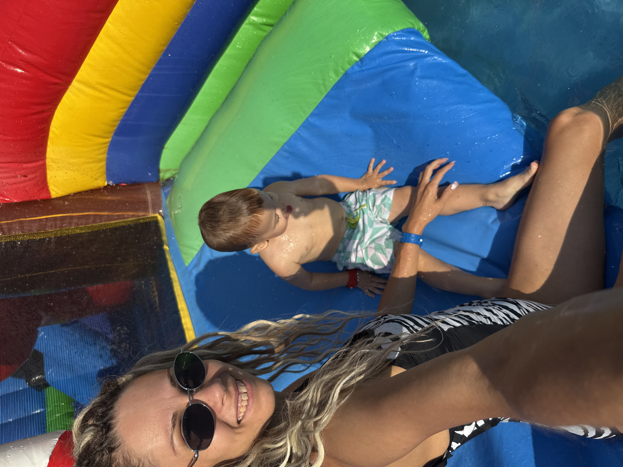 This media file shows two people, an adult woman and a young child, on a wet, inflatable water slide. The slide has sections of red, yellow, blue, and green. The woman is in the foreground, taking a selfie. She has long, blonde, curly hair and is wearing sunglasses and a black and white striped swimsuit. She is smiling and appears happy. The child, a boy, is lying on the blue section of the slide in the background. He is wearing light-colored swim trunks with a geometric pattern and has wet hair. He is looking to the side and appears to be relaxed or perhaps slightly bored. The slide is wet, indicating it is being used as a water slide, possibly on a sunny day given the lighting and shadows. The overall scene suggests a fun, recreational activity, likely at a water park or a backyard setup. There is no visible text in the media file.