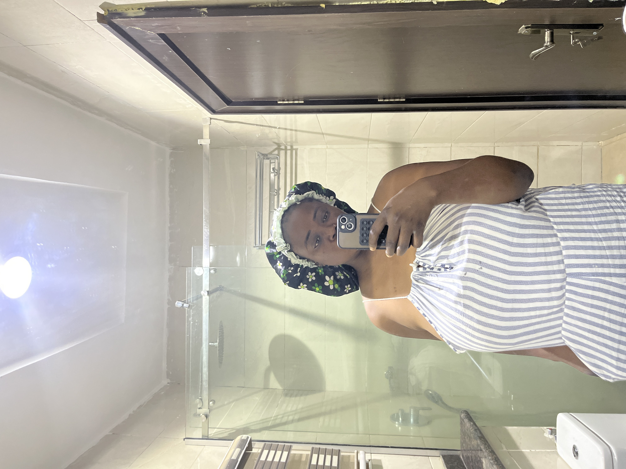 A woman in a floral hair bonnet and striped dress is taking a selfie in a bathroom. The bathroom has a glass-enclosed shower with a showerhead visible, tiled walls, a mirror, and bathroom fixtures. The location is likely in Abuja, Nigeria, but this is only inferred from the request.