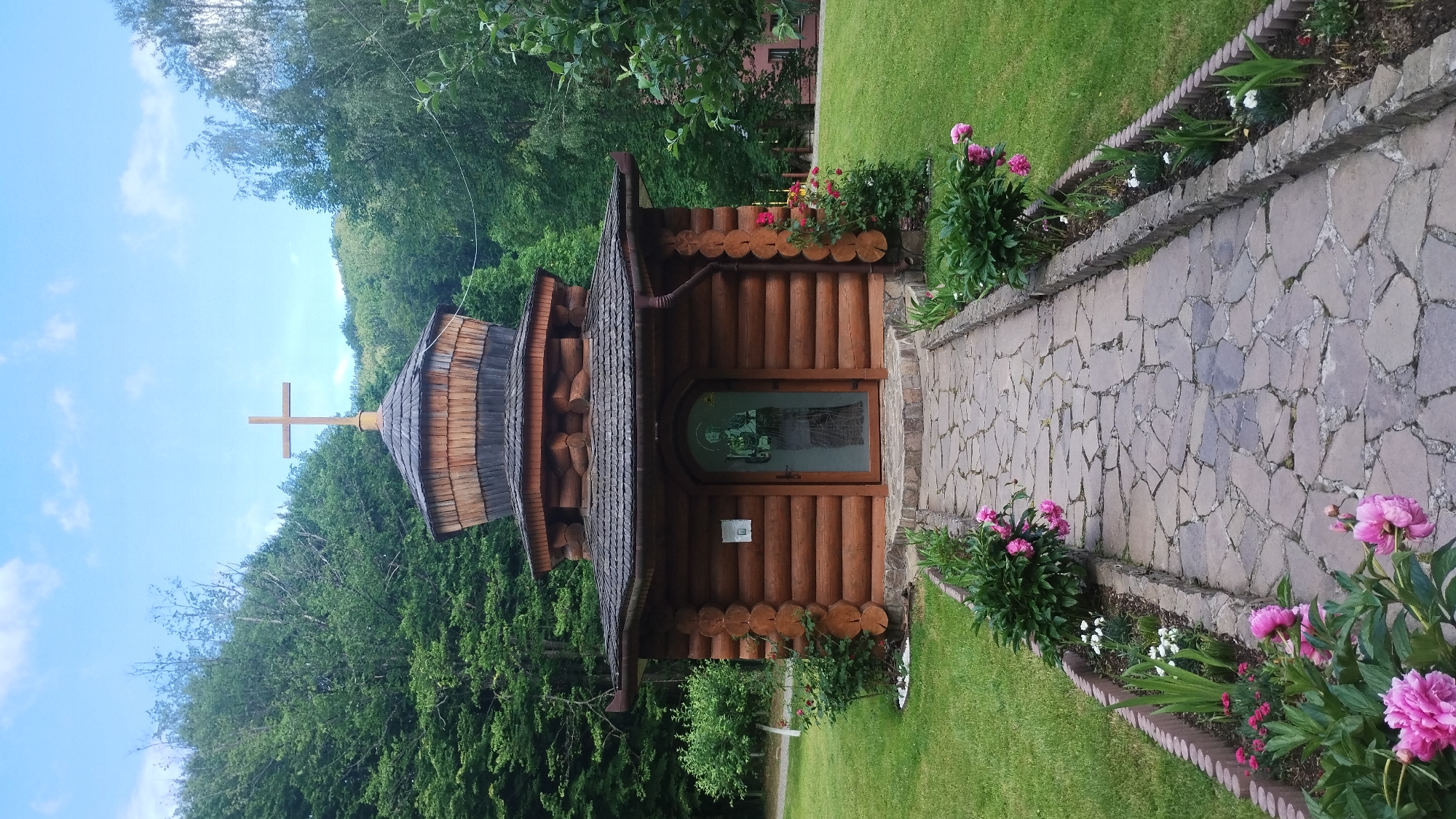 A frontal medium shot captures a small, wooden, log-built chapel with a domed roof, topped by a cross, in Syniak, Ukraine. The chapel has a glass door with a stained-glass image of a religious figure. A flagstone pathway leads to the entrance, flanked by flowerbeds with pink peonies and other small blooms. The chapel is situated on a grassy area surrounded by lush green trees under a partly cloudy sky. To the right of the chapel, a section of a building with a red roof is visible.