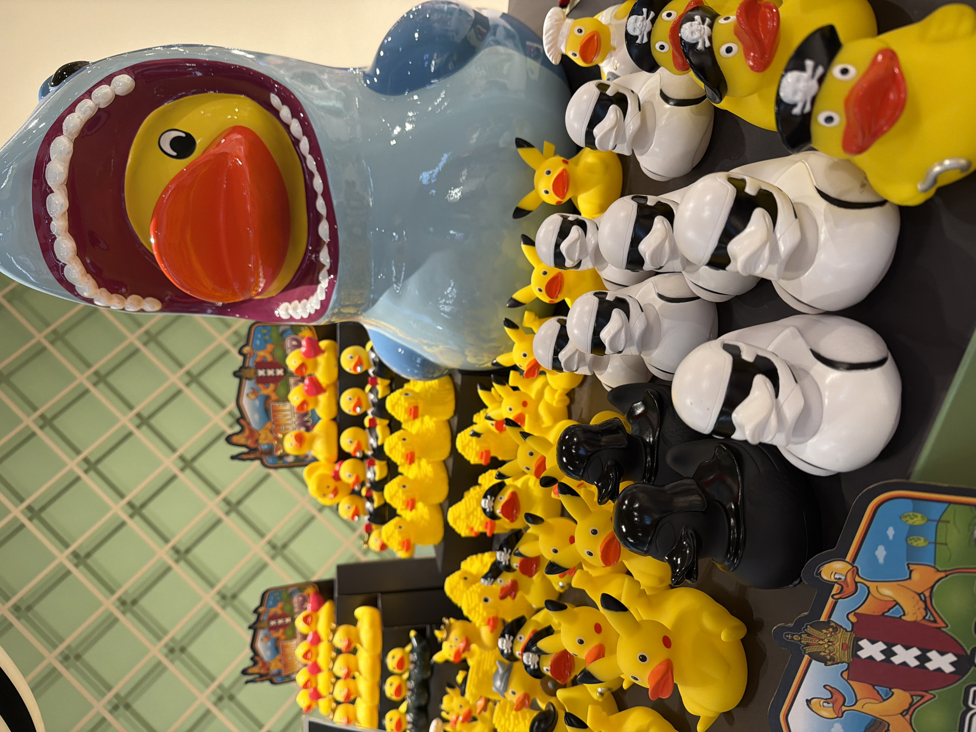 This media file features a variety of rubber ducks and Pikachu figurines displayed in a store setting. The main subject is a large, light blue shark-shaped object with an open mouth, revealing a yellow duck inside. Surrounding the shark are rows of rubber ducks, some yellow with pirate hats, and others white with black stripes. Numerous yellow Pikachu figurines are also visible, arranged in rows. In the background, a wall with a green grid pattern is partially visible. The objects appear to be for sale or display, likely in a novelty shop or toy store. The lighting suggests an indoor environment. There is no visible text in the main portion of the image, though two colorful signs with cartoon ducks and what appears to be a coat of arms with "XXX" are present at the bottom right. The overall scene evokes a playful and whimsical atmosphere.