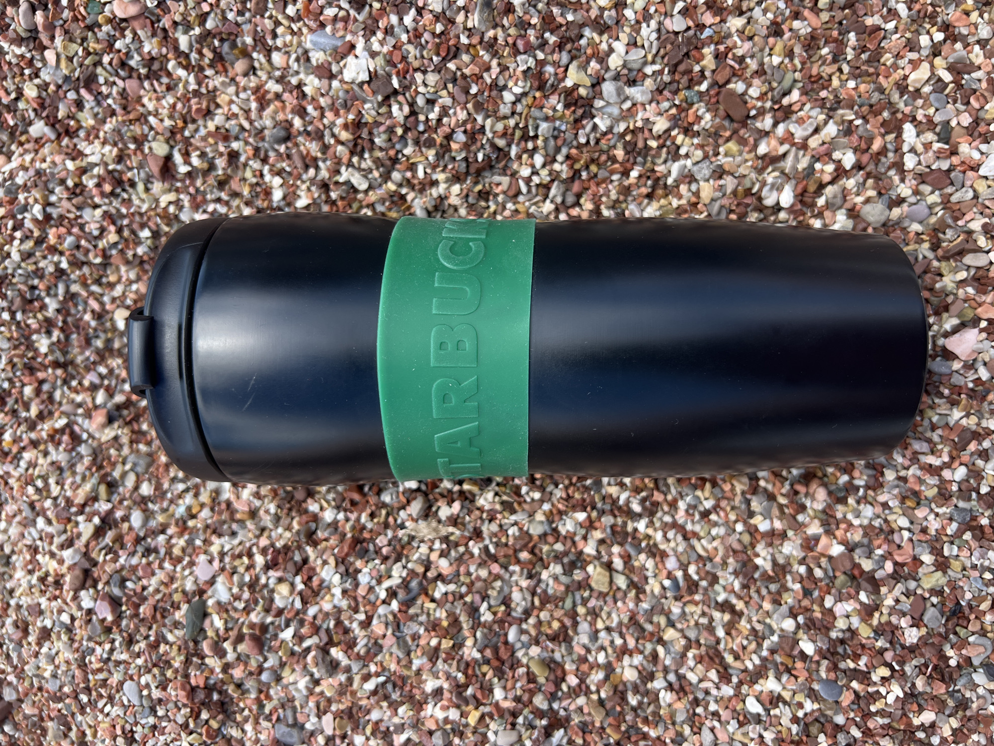 The image shows a black Starbucks tumbler lying horizontally on a surface covered with small, multicolored pebbles. The tumbler has a green band around it with the Starbucks logo embossed. The location is Petrovac centar, Montenegro, suggested by the pebble composition typically found on beaches in that region.