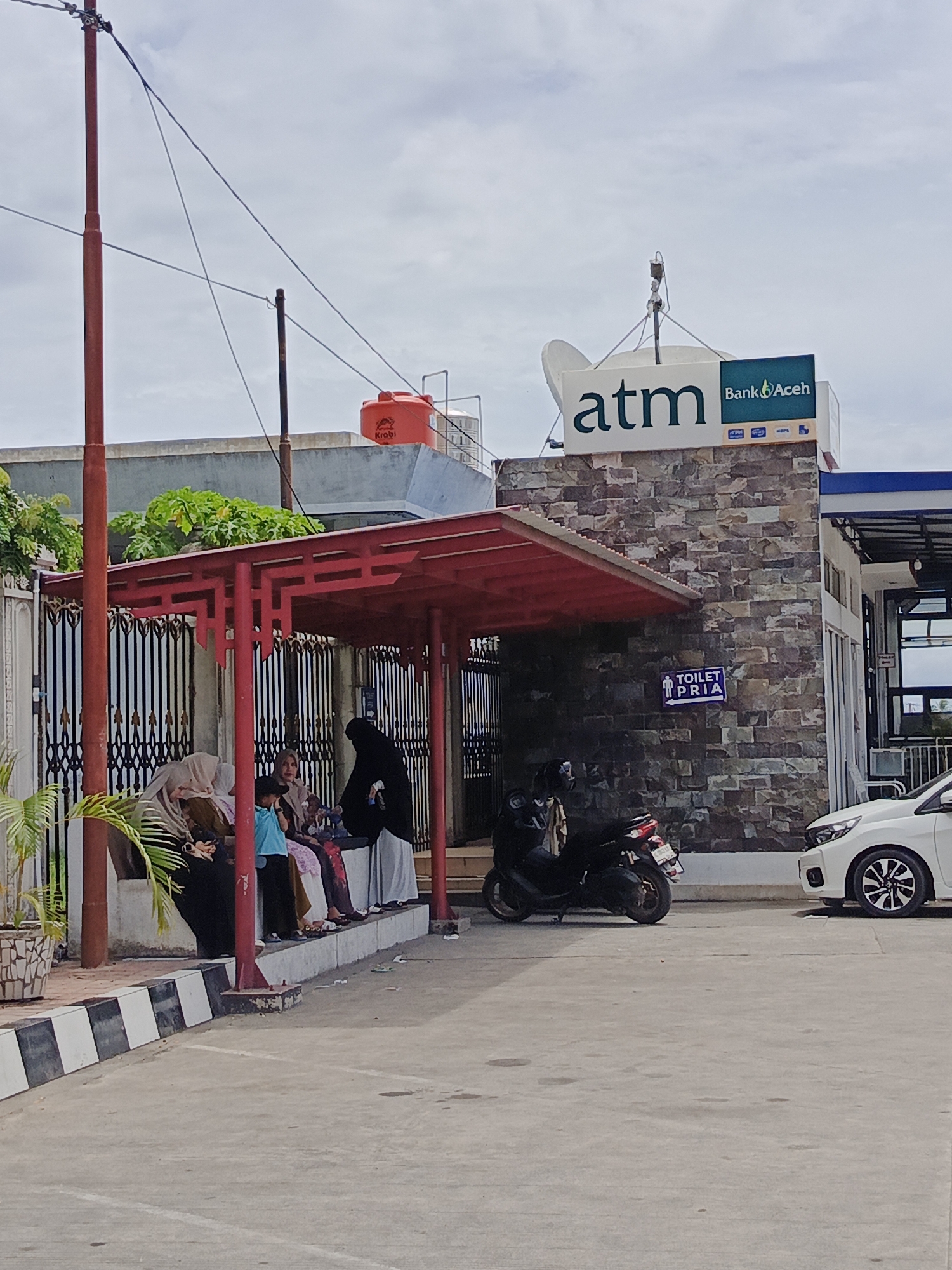 In Cotbadatunong, Indonesia, this image captures a scene outside a Bank Aceh ATM facility. A red awning supported by dark posts covers a waiting area where several individuals are seated on a bench. Among them are women wearing hijabs, a child in a blue shirt, and another person in a black hijab. Two motorcycles are parked nearby, and a white car is visible to the right. The ATM sign, featuring the Bank Aceh logo, is prominently displayed on the building, which is constructed with stone accents. A sign indicating "TOILET PRIA" (Men's Toilet) is mounted on the wall below the ATM sign. The environment appears to be an outdoor public space with a concrete ground surface. Overhead, power lines crisscross against a cloudy sky, and a large orange water tank is visible on the roof of an adjacent structure.