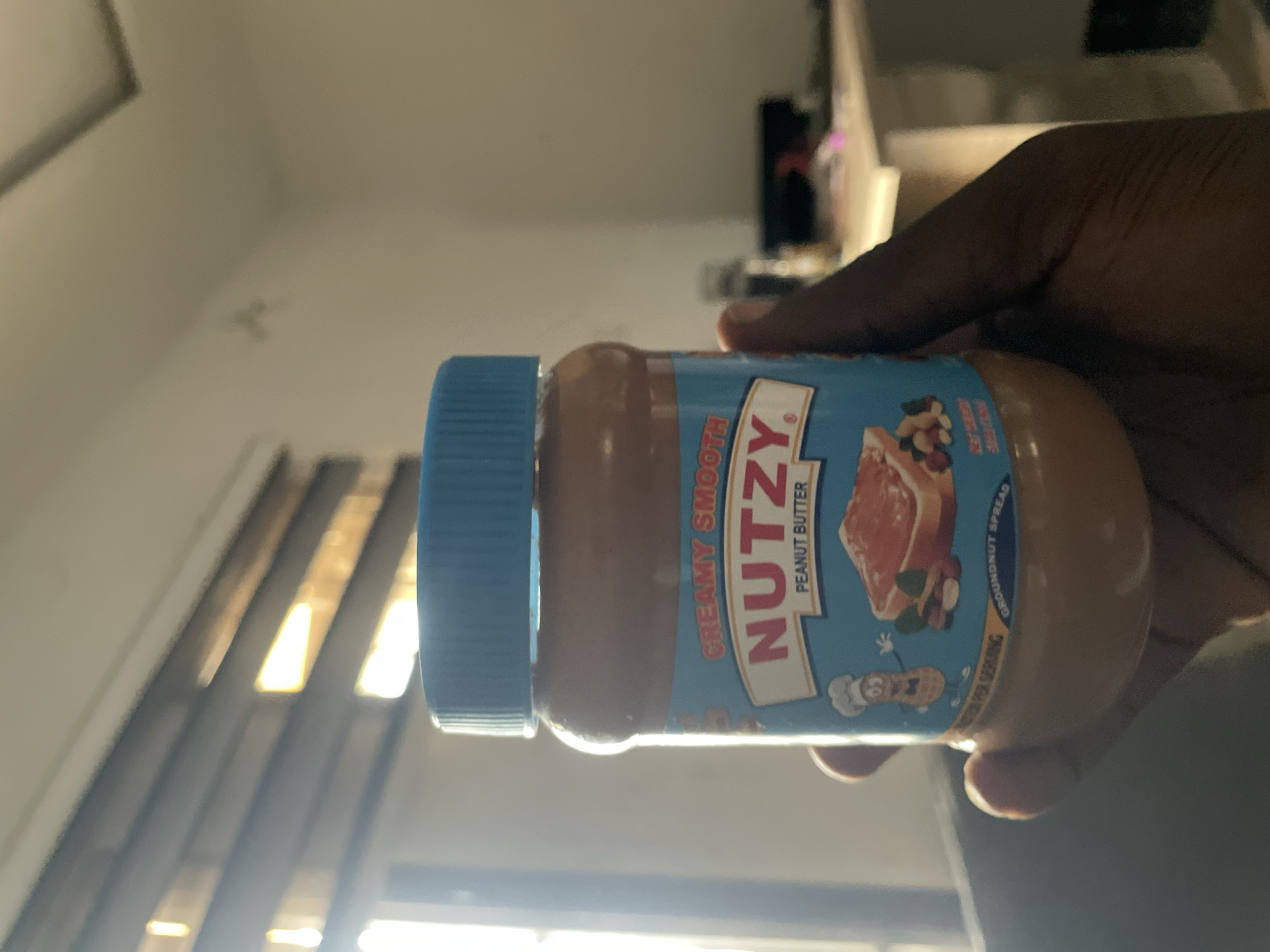 A close-up shot shows a person's hand holding a jar of "NUTZY" peanut butter with a blue lid. The jar's label is predominantly blue and features an illustration of a piece of toast with peanut butter, peanuts, and a cartoon peanut character dressed as a chef. The background is blurred but suggests an indoor environment with a window covered by blinds, indicating natural light entering the room. The overall setting appears to be domestic, potentially a home in Abuja, Nigeria, given the implied context.