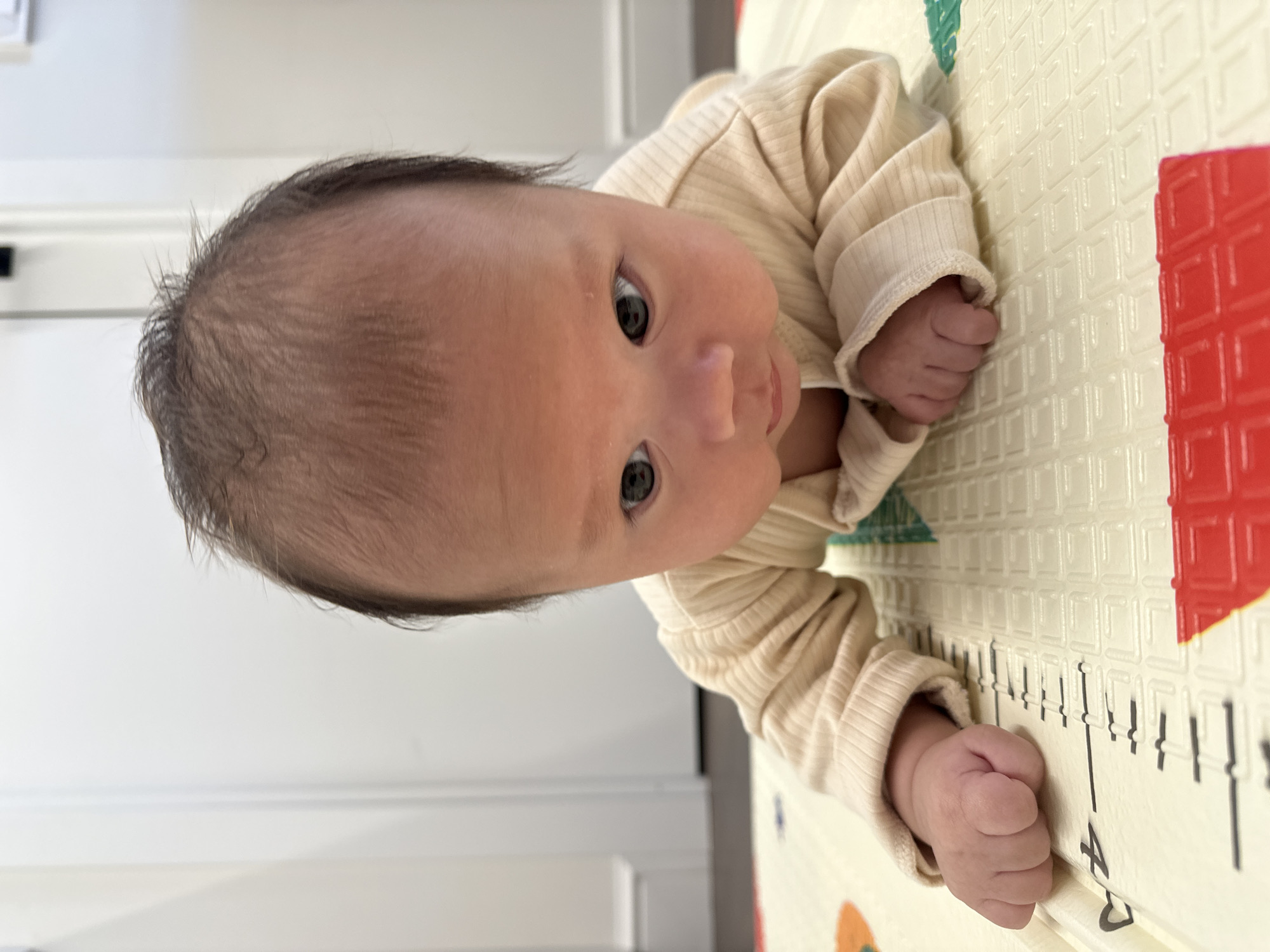 A baby is lying on their stomach on a play mat with a height chart. The baby is wearing a light tan, ribbed long-sleeved shirt. The baby has dark hair and light skin, with eyes looking up. The background includes a white wall. The scene is indoors. The location and city are unknown.