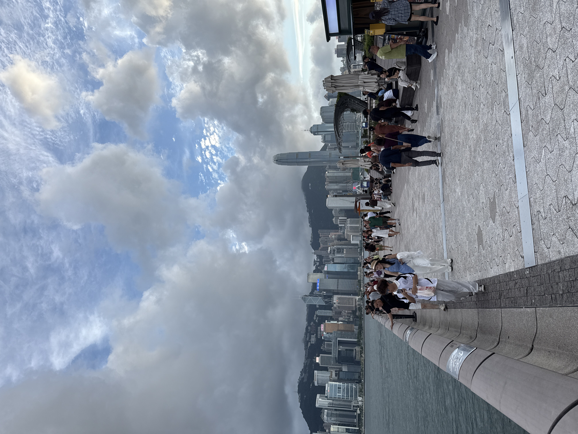 The image shows a daytime view of the Hong Kong skyline with a crowd of people along a waterfront promenade. The sky is partially cloudy, with patches of blue. The city skyline includes numerous skyscrapers, one notably taller than the others, with a mountain range as a backdrop. People are standing, walking, and sitting along the promenade, engaging in various activities such as taking photos and sightseeing. The promenade is paved with gray stone. Water can be seen at the bottom of the frame. The scene is likely taken from the Avenue of Stars in Tsim Sha Tsui, Hong Kong.
