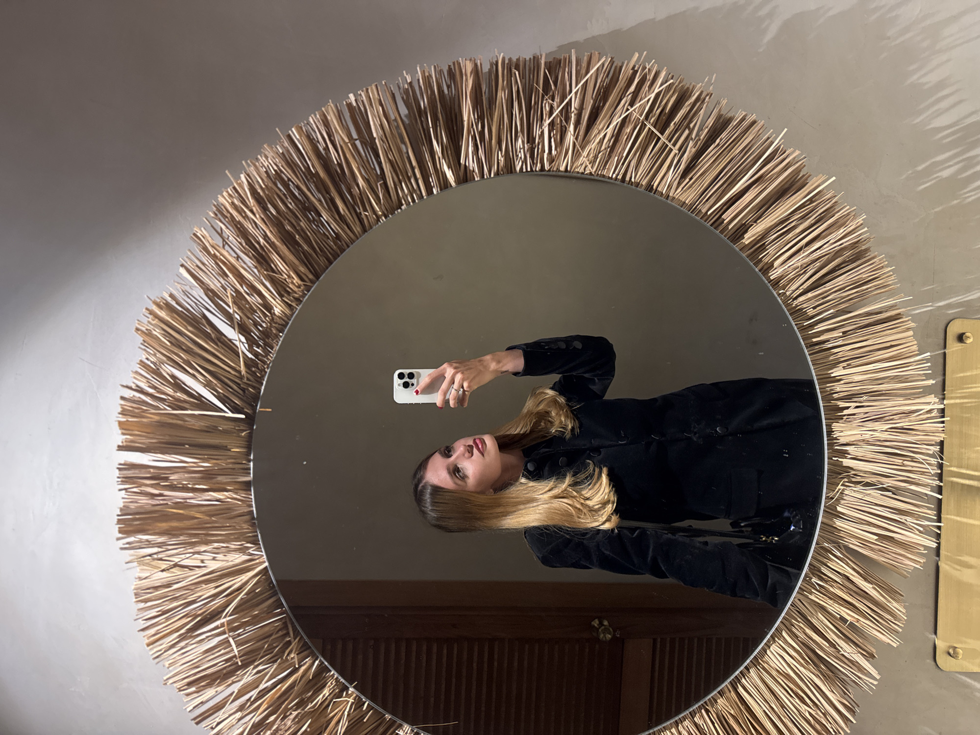 A woman is taking a selfie in a round mirror with a straw-like frame. The woman has blonde hair and is wearing a black jacket. The background includes a wall, and a door appears to be visible in the reflection. The location is presumed to be in Parakar, Armenia.