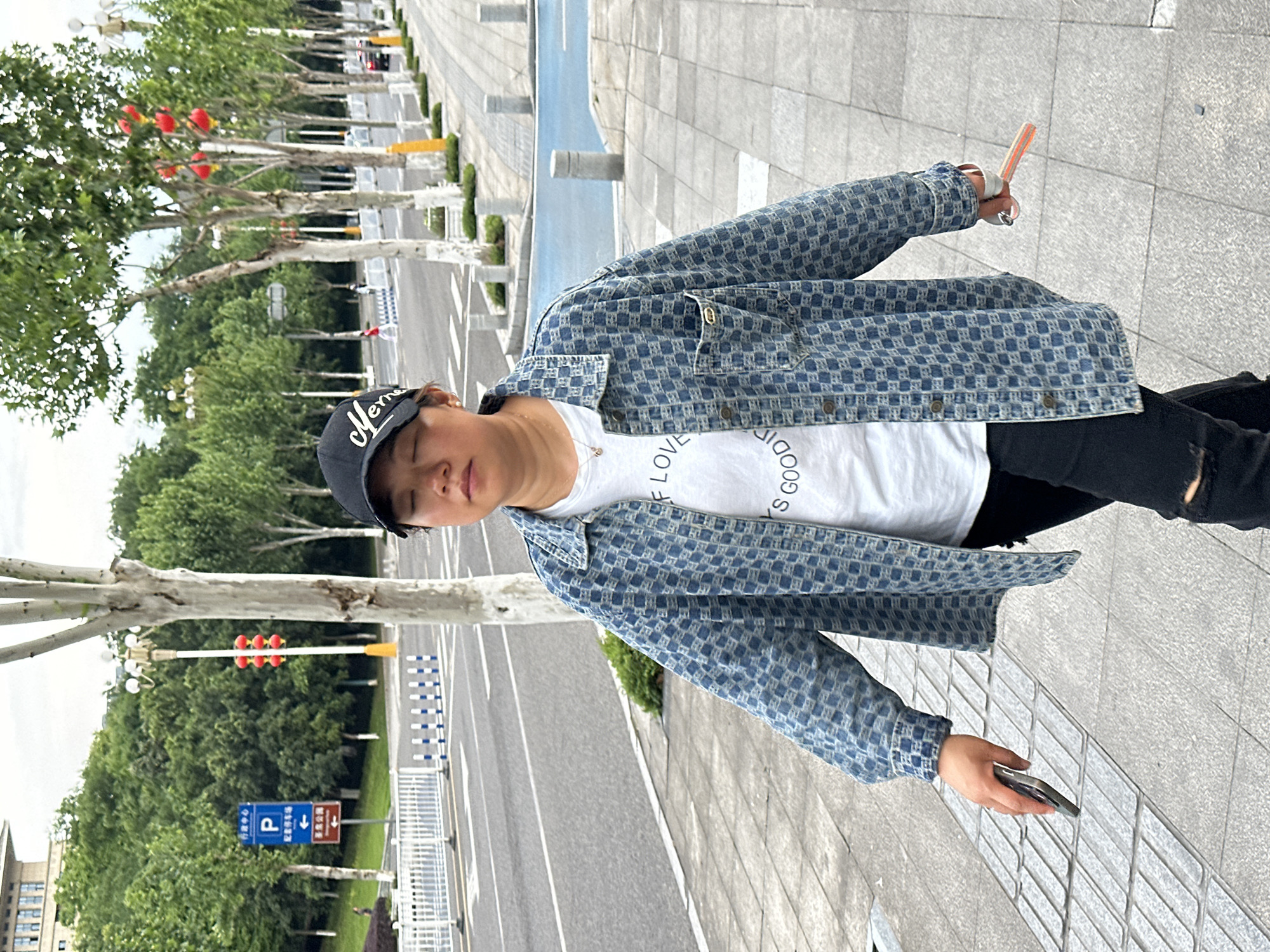 A person is depicted standing on a grey tiled sidewalk next to a road, looking downwards with their eyes closed or nearly closed. They are wearing a black baseball cap with white text, a white t-shirt with partially visible black text, a blue patterned denim jacket, and black pants. In their left hand, they hold a smartphone, and in their right hand, they hold what appears to be a set of keys or a small item attached to a strap.
The road in the background is lined with deciduous trees featuring light-colored bark and green leaves. Several red lanterns are suspended from the branches of the trees on the left side of the road. Further along the street, light poles, traffic lights (displaying red), and road barriers are visible. Road signs with Chinese text provide location cues: a blue sign with a white 'P' symbol indicating parking and the characters "行政中心" (Administrative Center), and another sign below it reading "P 机动车停车场" (Motor Vehicle Parking Lot) with an arrow. A brown sign also points towards "景区方向" (Scenic Area Direction).
The scene is set during the daytime under an overcast sky, providing soft, diffused lighting. The text on the black cap appears to be "Meme" or "Merma". The white t-shirt has "LOVE GOOD" and part of an 'S' visible, possibly from words like "GOODS" or "GOODIES". The person's posture and closed eyes suggest a moment of relaxation or contemplation in a bustling street environment. The visible Chinese signage confirms the location as being in China, specifically Jiangjin as per context.