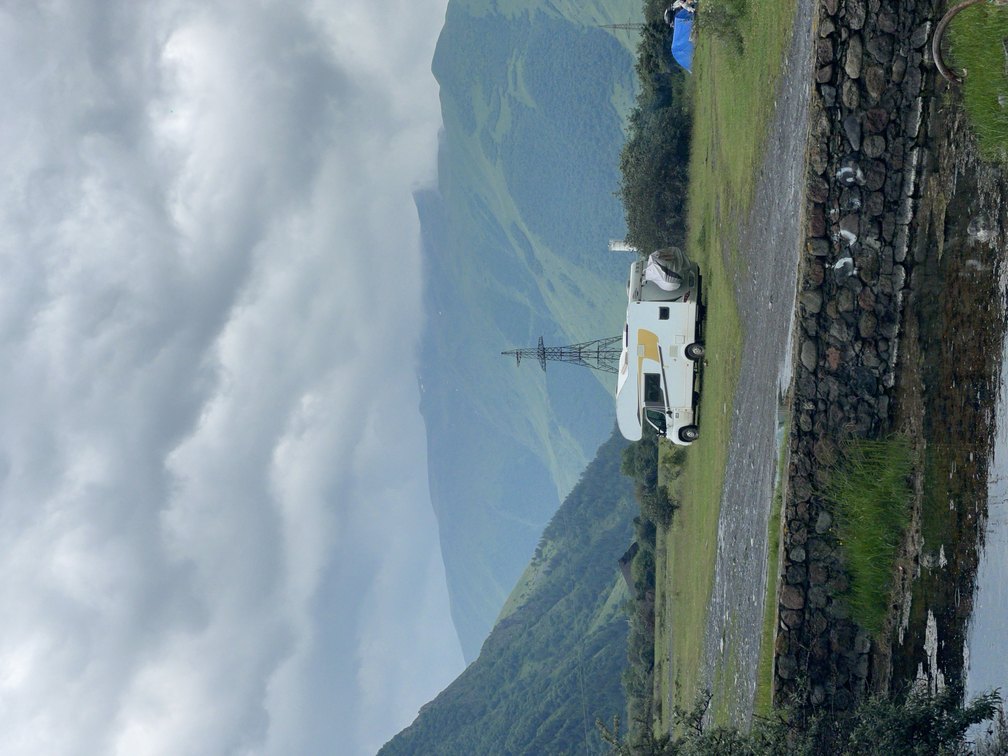 The image depicts a campsite in a mountainous region, possibly near Stepantsminda, Georgia, under a cloudy sky. In the foreground, a stone wall runs horizontally, with a small body of water to its left and grass to its right. Beyond the wall, a flat, grassy field stretches towards the background. A white camper van is parked in the middle ground of the field, facing towards the left. Behind the camper van, there is a blue tent pitched, with a couple of figures partially visible near it. Further in the background, several green mountains rise, with a tall electrical tower standing prominently on the left side of the frame and another smaller one on the right. The overall scene suggests a remote, natural setting with evidence of outdoor recreational activity.