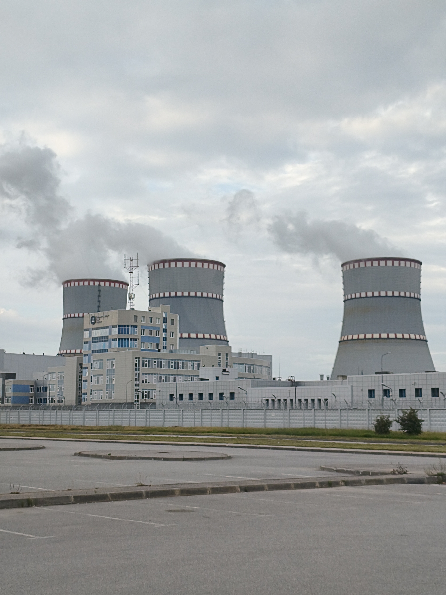 This is a long shot, daytime, outdoor, eye-level, full shot of the Leningrad Nuclear Power Plant in Sosnoviy Bor, Russia. The image is captured on an overcast day with a sky full of thick, gray clouds. In the foreground, there is an empty asphalt parking lot with marked spaces and concrete dividers. Beyond the parking lot, a chain-link fence with barbed wire runs across the frame, separating the lot from a grassy area. In the middle ground, there are several low-rise buildings with a modern architectural style, featuring white and blue facades and many windows. Some of these buildings have antennae on their roofs. Behind the buildings and to the left and right, three massive cooling towers dominate the scene. The cooling towers are cylindrical, gray structures with red and white bands around their upper rims. Thick white plumes of steam are billowing from the tops of the two cooling towers on the left, indicating active operation. The cooling tower on the far right also has a visible plume of steam. The overall impression is one of an industrial complex under a muted sky.