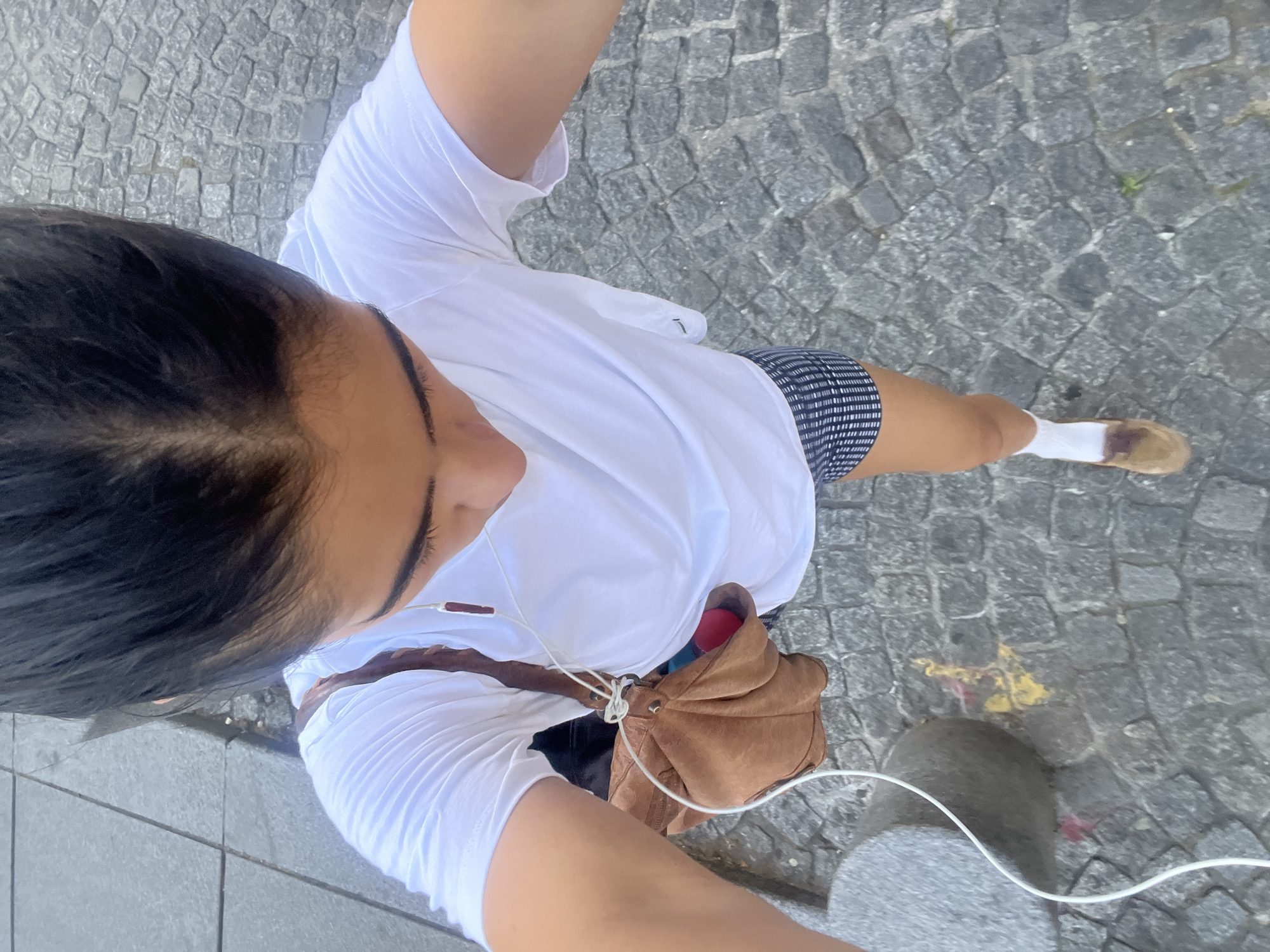 A high-angle photograph captures a person, presumed to be female, standing on a cobblestone surface. The individual is wearing a white t-shirt, patterned shorts, white socks, and tan shoes. A brown leather bag hangs across their torso. They are also wearing earbuds with white wires. The cobblestone surface indicates a pedestrian area, potentially a street or square in Paris, France. A concrete post or bollard is visible to the right of the individual.