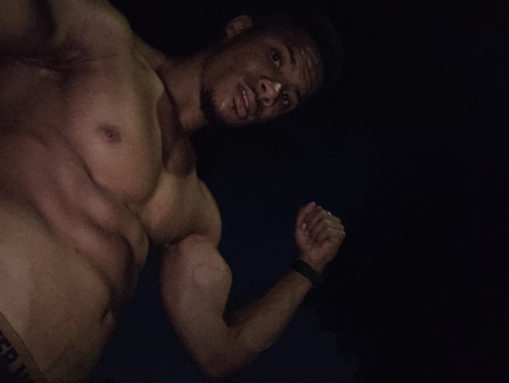 The image is a close-up, low-angle, outdoor photograph of a muscular man flexing his bicep. The man is shirtless and appears to be of African descent. He has a short beard and short hair. He is looking upwards and to his right, with sweat on his forehead and nose. He is wearing dark-colored briefs, the waistband of which is partially visible. The background is entirely black, indicating a dark environment, possibly nighttime. The lighting is focused on the man's upper body, highlighting his musculature. There is no indication of the city of Varna, Bulgaria, or any specific location.