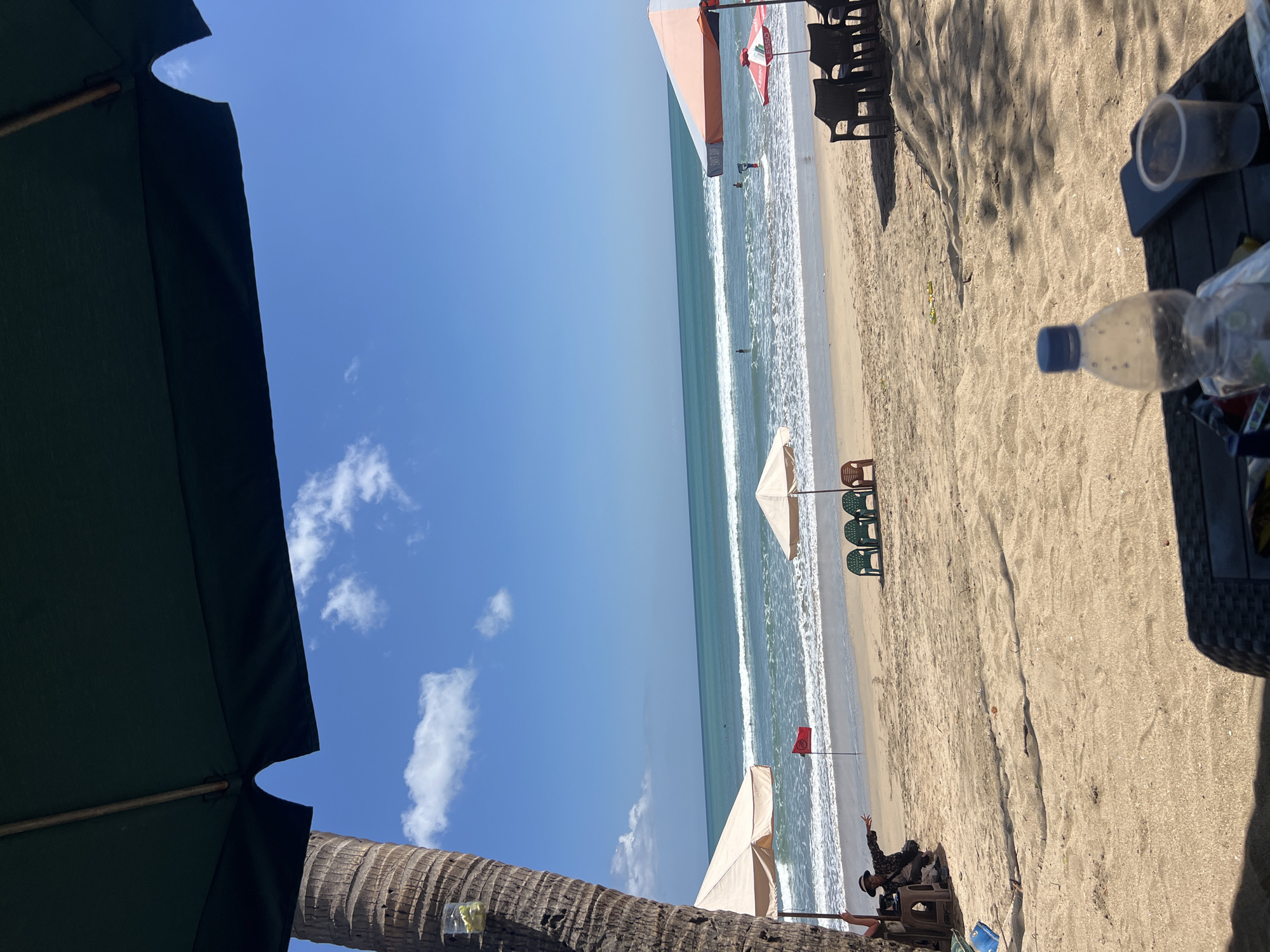 A bright, sunny day at Kuta Beach, Indonesia, as indicated by the clear blue sky with scattered white clouds, the gentle waves of the ocean, and the palm tree framing the left side of the image. In the foreground, a person sits under a large dark umbrella, pointing towards the sea. Beach chairs and umbrellas are set up on the sand, with a red flag near the water's edge, suggesting a designated swimming area or a warning. Several people are visible in the water, enjoying the waves. The scene captures a typical day at a popular beach destination.