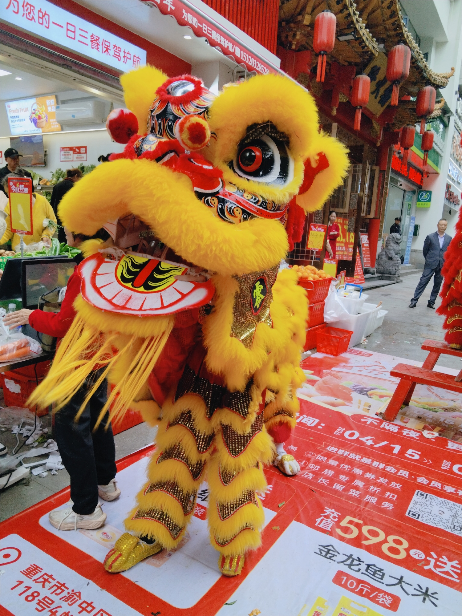 A full shot reveals a vibrant scene featuring a traditional Chinese lion dance costume performer outdoors. The lion is predominantly yellow with red and black accents, including gold embellishments on its legs and a decorative fringe. The performer is partially visible behind the lion's head, wearing black pants and white shoes.
The lion stands on a red promotional advertisement printed on a white background, which is laid out on the ground. The advertisement features large Chinese characters and QR codes.
In the background, a commercial area is visible with stores, red lanterns hanging overhead, and pedestrians. To the left, people are shopping at a produce stand with fruits and vegetables. To the right, a man in a suit stands on the sidewalk. The location is identified as the Yuzhong District, Chongqing City, China, based on the text on the advertisement.