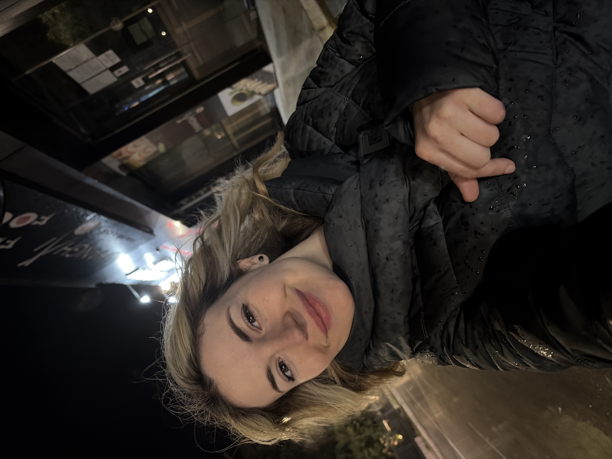 A woman with blonde hair smiles at the camera, wearing a dark, wet, puffy jacket. She holds her hand in a "hang loose" gesture, or "shaka sign." The background shows a dimly lit urban setting, possibly outdoors at night, with blurry lights and reflections suggesting it may be raining. The location appears to be in Budva, Montenegro, based on visual elements such as architectural style. The overall scene suggests an informal moment captured in a bustling urban environment during or after a rainfall.