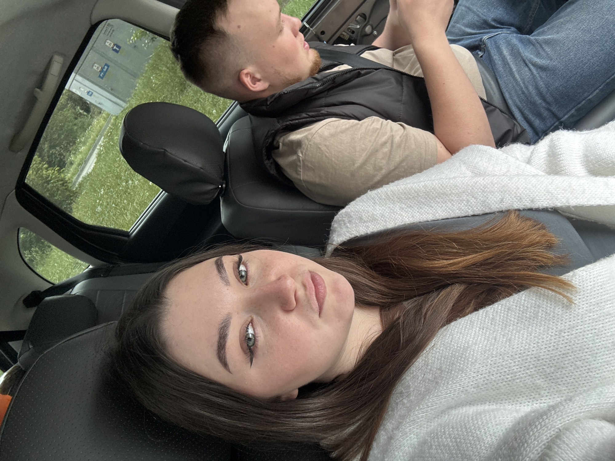 Two people are inside a car. A woman with long brown hair and wearing a white sweater is lying down on the back seats. A man is seated in the front passenger seat, wearing a black vest over a beige shirt and blue jeans. He is looking at a cell phone. Outside the car window, there is green vegetation and street signs. The location is Perm, Russia.