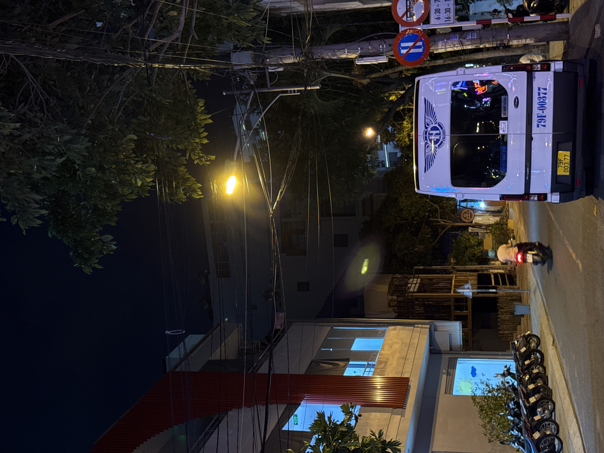 This is a nighttime street scene in Nha Trang, Vietnam. A white van with the license plate "79F-00377" is parked on the right side of the road, facing away from the camera. The van has a logo with wings and the letter "H" on its rear window. Next to the van, a utility pole is covered in numerous electrical wires. Two circular traffic signs are visible on the pole; one depicts a blue circle with a white horizontal line and a red diagonal line, and the other displays "29 CHOP" and time information like "6:30 - 7:" and "16:30 - 1".
To the left of the van, a row of parked motorcycles is visible along the sidewalk. Further to the left, a building with a red facade and illuminated windows with a blue glow is present. A person is riding a scooter on the road, heading away from the camera and towards the van. Trees with foliage are spread throughout the scene, with some leaves illuminated by streetlights. The street is paved and appears to be in a relatively urban area.