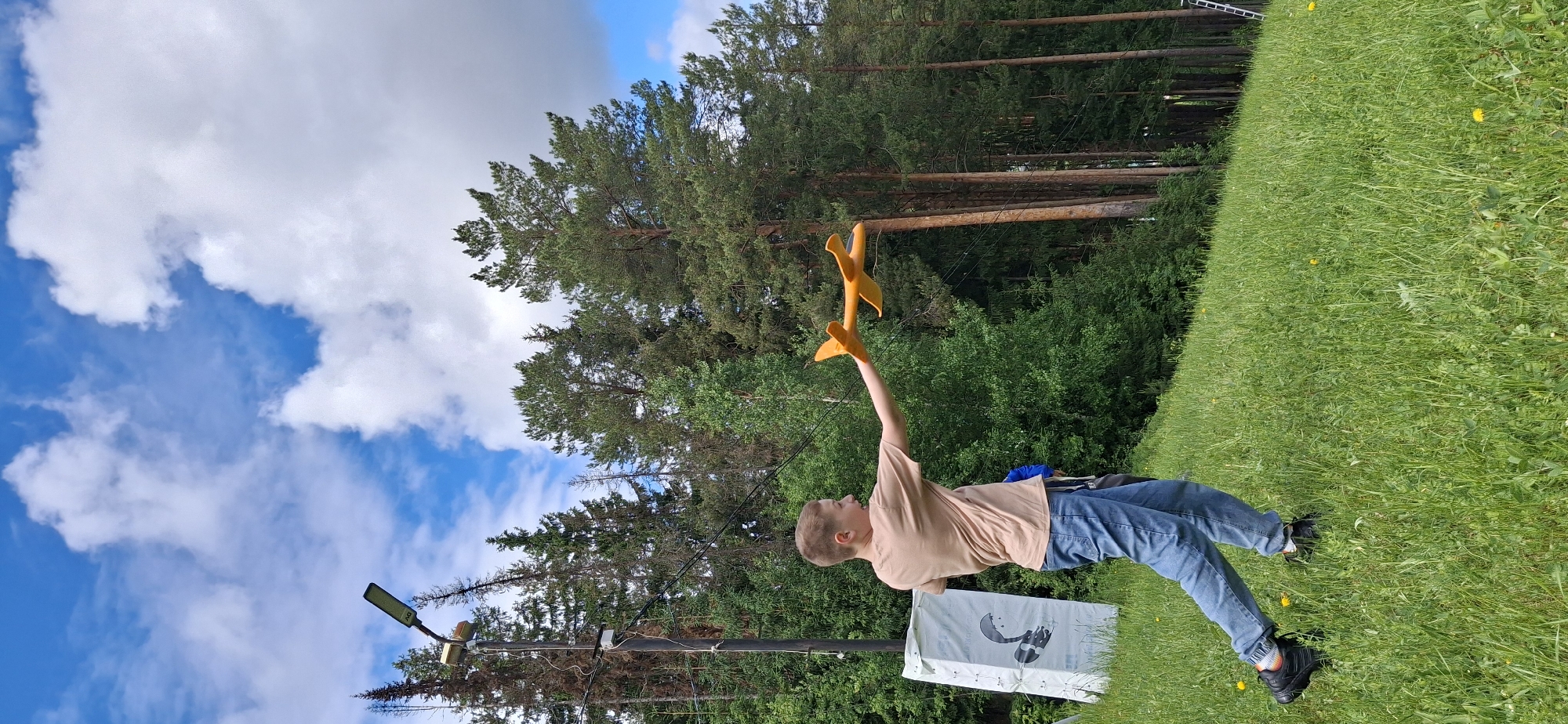 The image shows a young boy in an outdoor environment. The boy is wearing a beige t-shirt and blue jeans, and he is holding a yellow toy airplane above his head with his right arm. He is standing on a grassy hill. In the background, there are tall trees, bushes, and a blue sky with white clouds. There is also a street light pole on the left side of the frame and a white sign with a design on it near the boy. The location is Прикамский, Russia, based on contextual information.