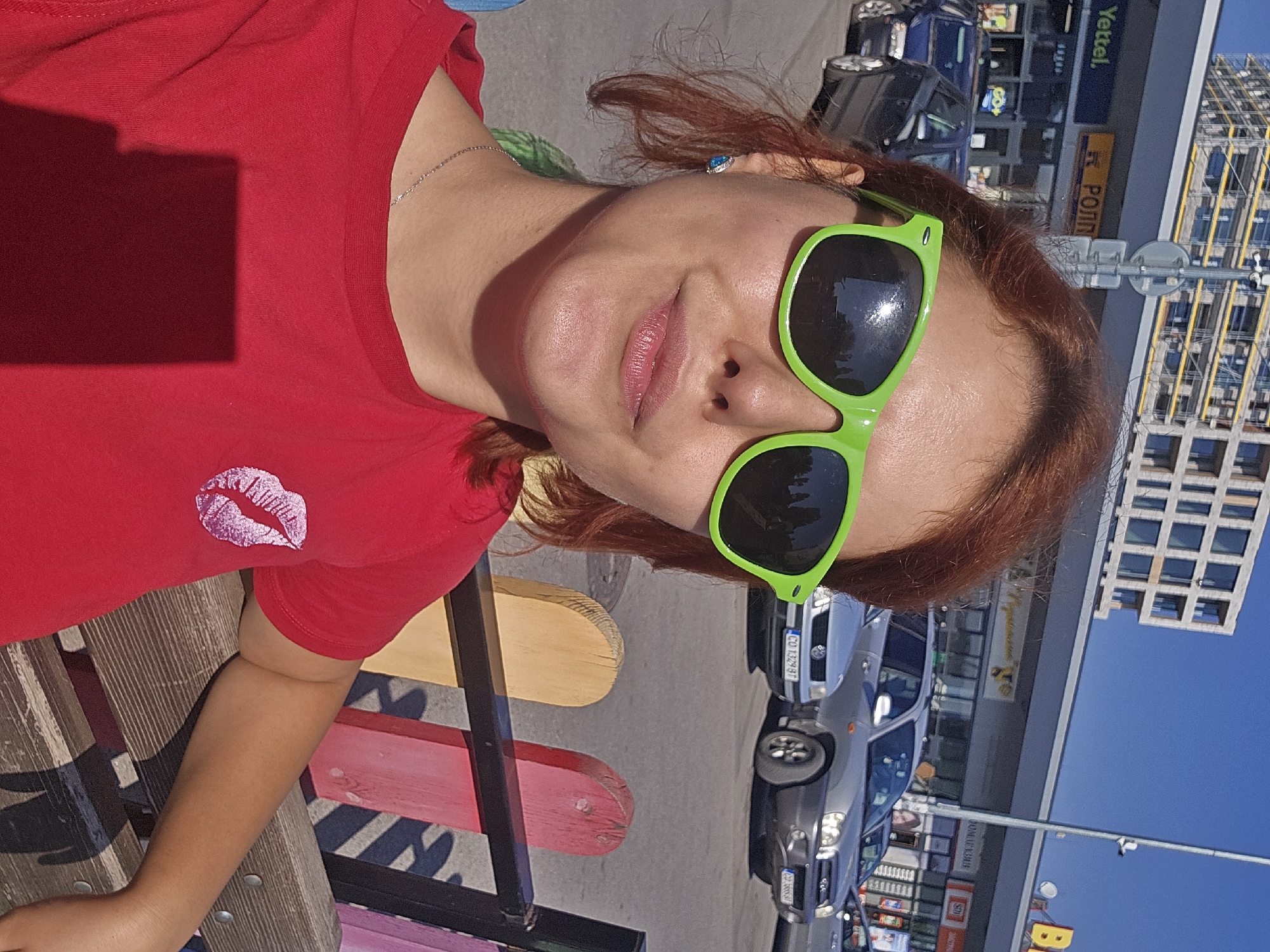 A young woman with auburn hair and bright green sunglasses is sitting on a bench outdoors. She is wearing a red t-shirt with a pink lipstick kiss graphic on the left breast and a silver necklace. She is looking at the camera with a slight smile. Behind her, cars are parked in front of a row of shops, and a multi-story building under construction is visible in the background. The sky is clear and blue.