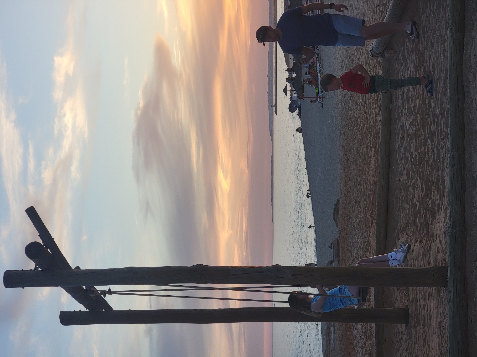 The image depicts a beach scene in Quarteira, Portugal during sunset. In the foreground, a wooden swing set is visible, with a young girl on the swing, wearing a blue top and shorts. To the right, a man in a blue t-shirt and shorts, wearing a baseball cap, is walking with a young boy in a red shirt and green pants. The beach is sandy, with people relaxing on lounge chairs in the background. The ocean stretches to the horizon, with a hazy sky displaying hues of orange, pink, and blue.