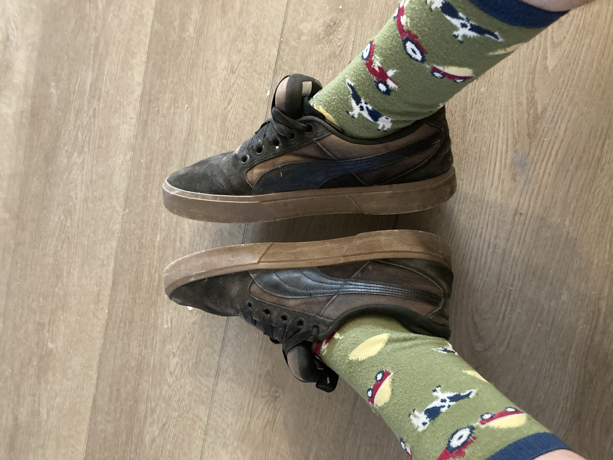 The image shows a pair of dark brown suede athletic shoes with brown rubber soles. The shoes are worn by a person whose feet are covered by green socks with a pattern of red cars and what appear to be abstract shapes. The person is standing on a light brown wooden floor. The scene is indoors, and there is no visible indication of the location being in Round Rock, United States of America, or any other specific city. The lighting suggests an overhead light source.