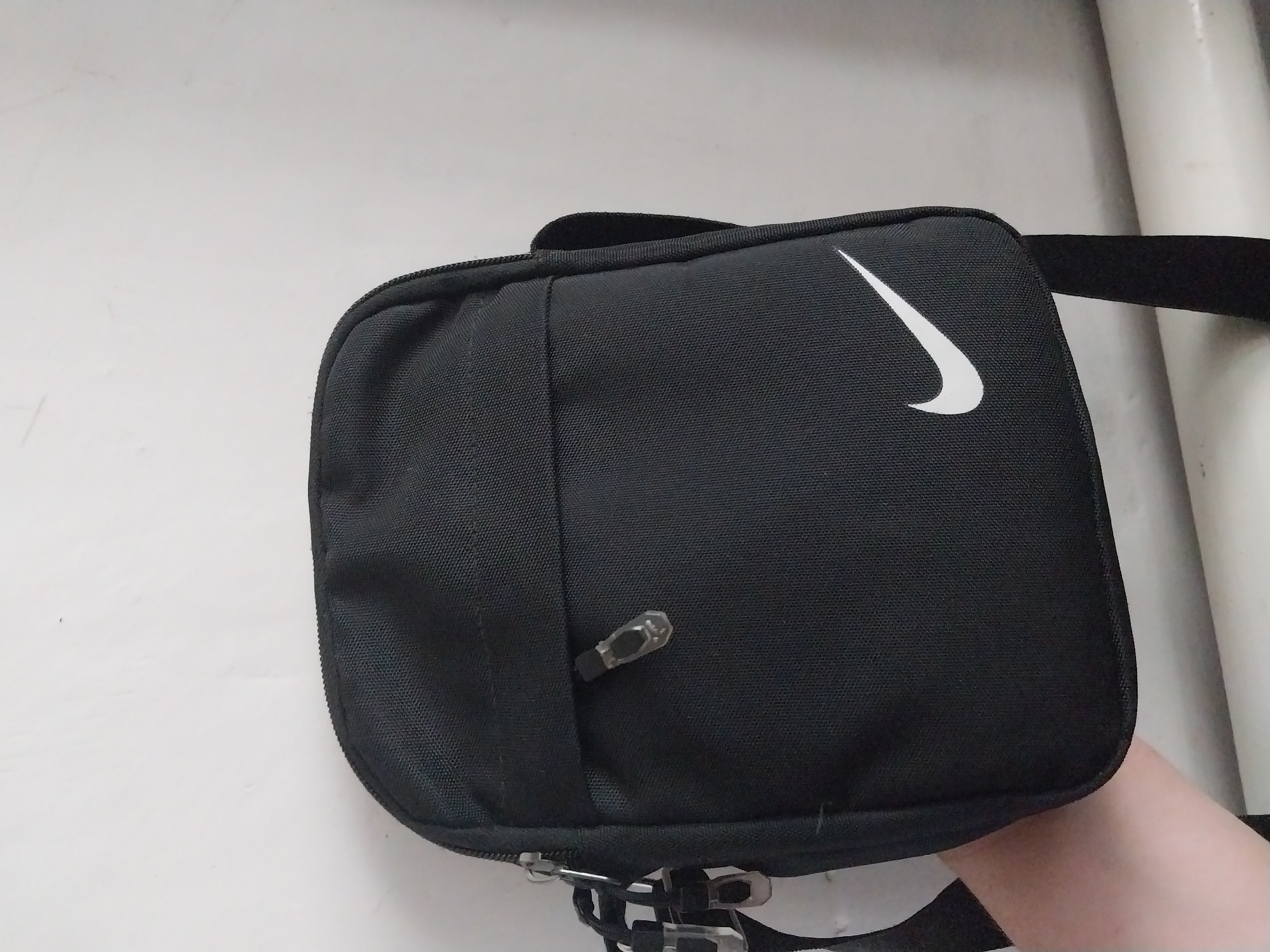 A black Nike crossbody bag is held by a person's arm against a white wall. The bag has a zipper closure on the top and a prominent white Nike swoosh logo on the front. The bag appears to be made of a durable, textured fabric. The background is a plain white wall, with some visible texture and possibly some faint smudges. The lighting is even and suggests an indoor environment. There are no specific identifiable landmarks or features that would indicate a location within the city of Ikove, Ukraine.