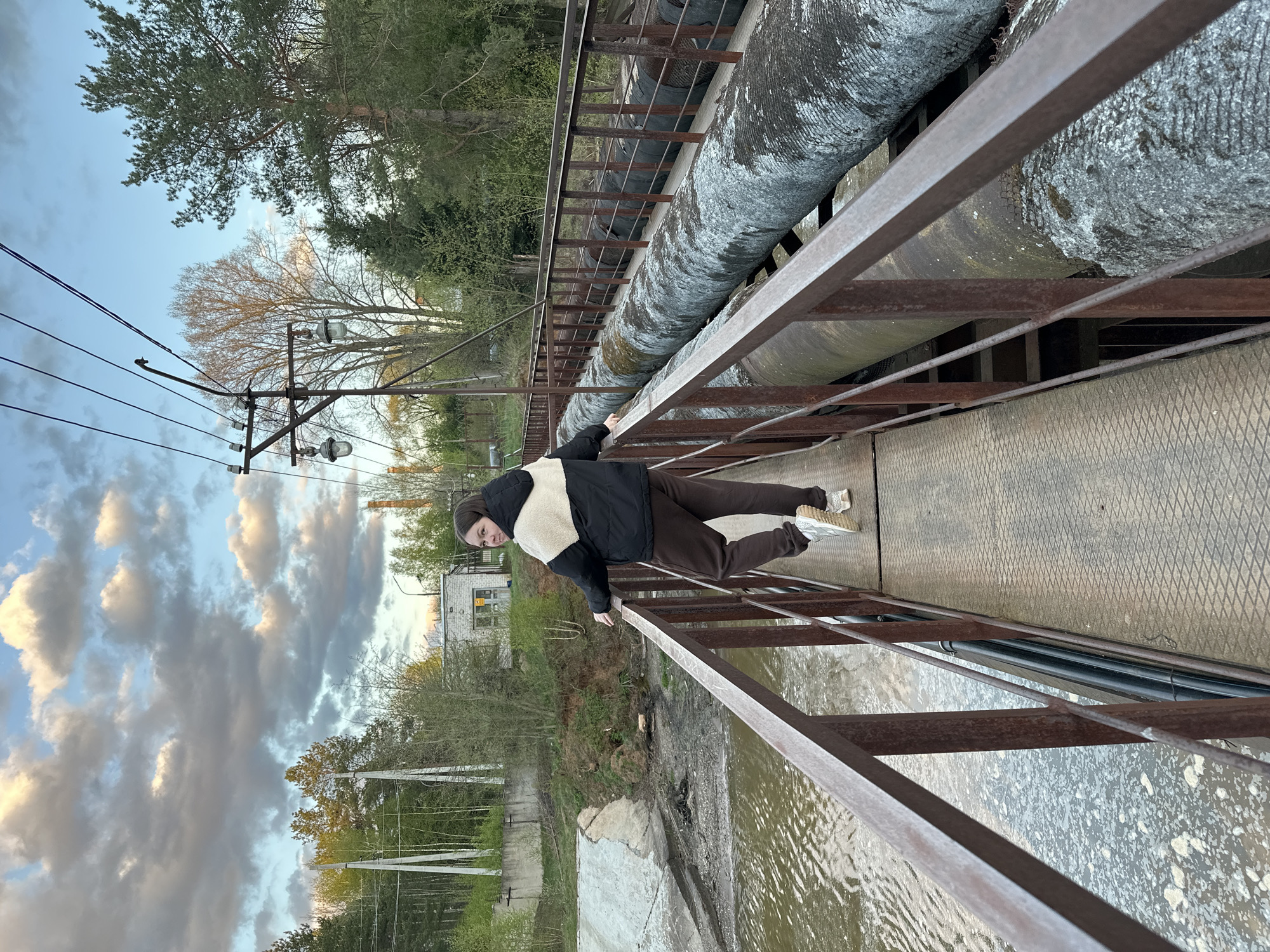 The image depicts an individual positioned on a metal bridge in Chaikovsky, Russia. The person is leaning against the railing, with a body angle that suggests a precarious stance. The person is wearing a black jacket with a white fuzzy interior, brown pants, and light-colored sneakers. The bridge spans over water, with a large pipe running along its side. The background includes trees, power lines, and a building in the distance. The sky is cloudy, indicating either morning or evening light conditions. The bridge is constructed of metal with a diamond pattern deck, and the railings are rusty.