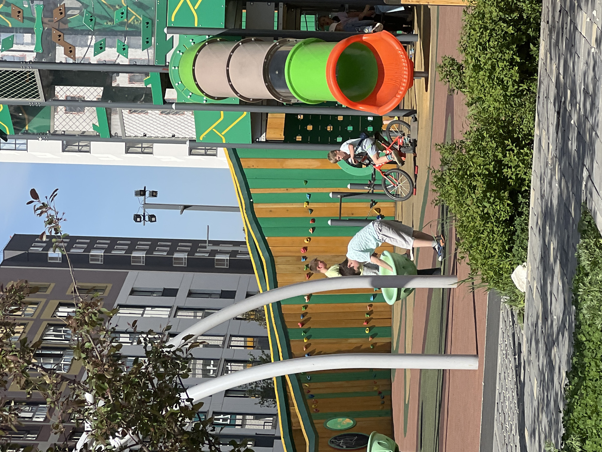 The image shows a playground located in Yekaterinburg, Russia. The playground is colorful with green, yellow, orange and brown elements. A tall apartment building is in the background. Several individuals are on the playground, including a child riding a red bicycle near the top of a tubular slide structure. Another child and an adult are near a rock-climbing wall with green climbing holds. The play area is adjacent to a paved area and dense green foliage.