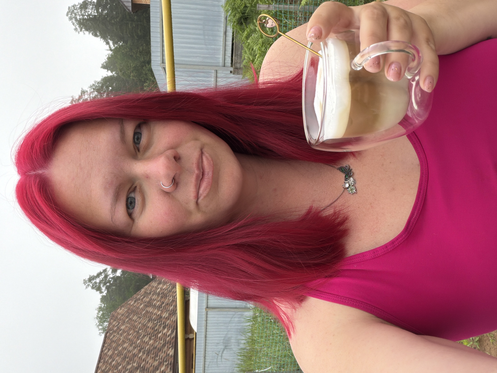 The image, presented in a tilted orientation, depicts a woman taking a selfie outdoors, likely in Chaikovsky, Russia. She has vibrant, bright red hair, light blue eyes, and a silver hoop nose ring. Her gaze is directed at the camera, and she wears a subtle, calm expression with a hint of a smile.
She is dressed in a vivid fuchsia-colored tank top and a silver necklace adorned with a small, intricate pendant that appears to be a butterfly or floral design. In her right hand, she holds a clear glass mug containing a layered beverage, resembling a latte or cappuccino, complete with a thick layer of white foam on top. A slender, golden stirrer featuring a delicate heart-shaped embellishment extends from the drink. Her fingernails are manicured with a light, possibly glittery, polish.
The background suggests an overcast daytime setting. Visible elements include dark green trees and foliage, a portion of a building with a textured, possibly tiled, roof, and a light-colored corrugated metal structure or fence. These details indicate a residential or backyard environment. There is no visible text in the image.