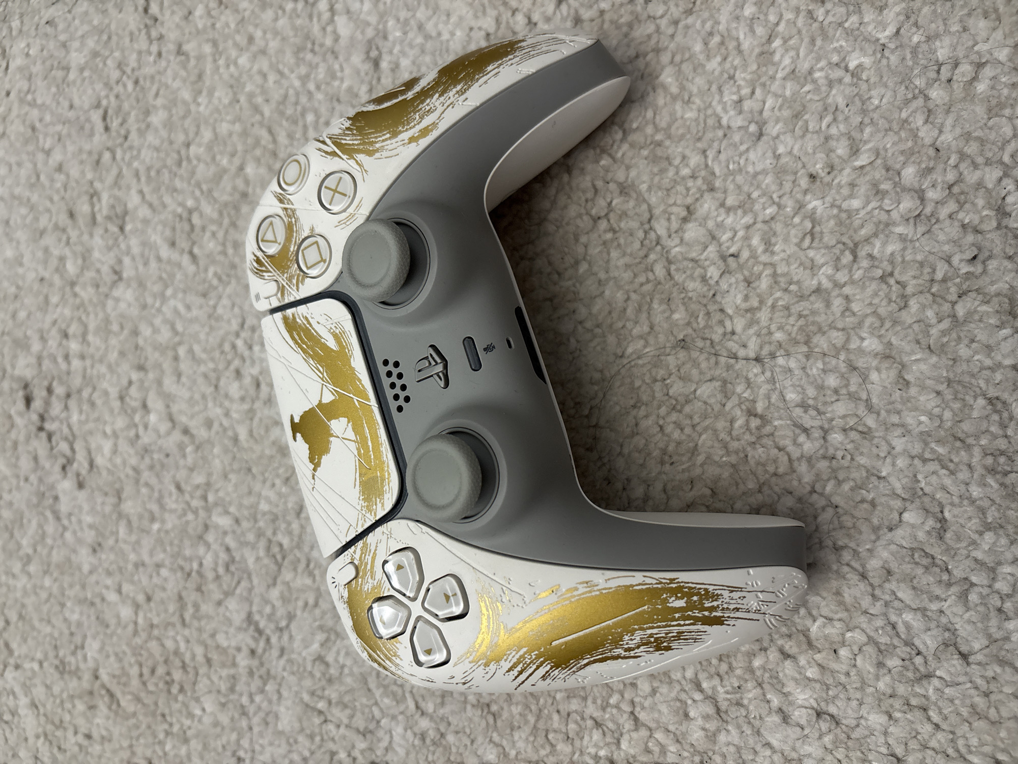 A white and grey PlayStation 5 DualSense controller with gold brush stroke style decoration is positioned on a light grey carpet. The controller is oriented diagonally with the left side facing towards the bottom of the frame. The carpet appears to be short pile and slightly textured. The location is Unknown, Unknown.