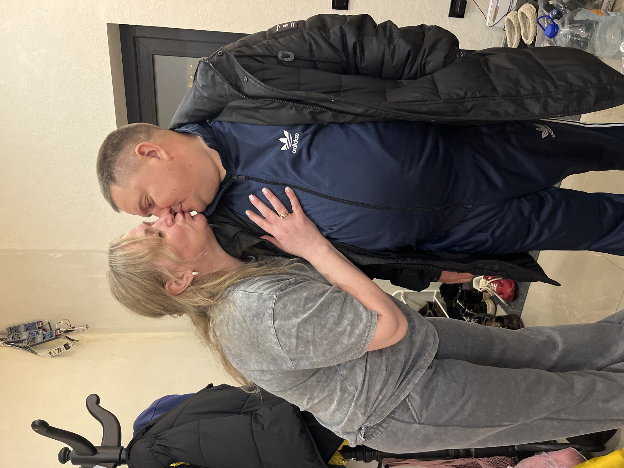 A man and woman are kissing indoors, possibly in a residence in Новая Чернушка, Russia. The man is wearing a dark blue Adidas tracksuit with a black puffer jacket over it. The woman is wearing a gray t-shirt and gray sweatpants. The background includes a doorway, a coat rack with a black jacket, and miscellaneous items on the floor and in the corner of the room. There appears to be electrical wiring exposed on the wall near the coat rack. The woman is wearing a ring on her left hand.