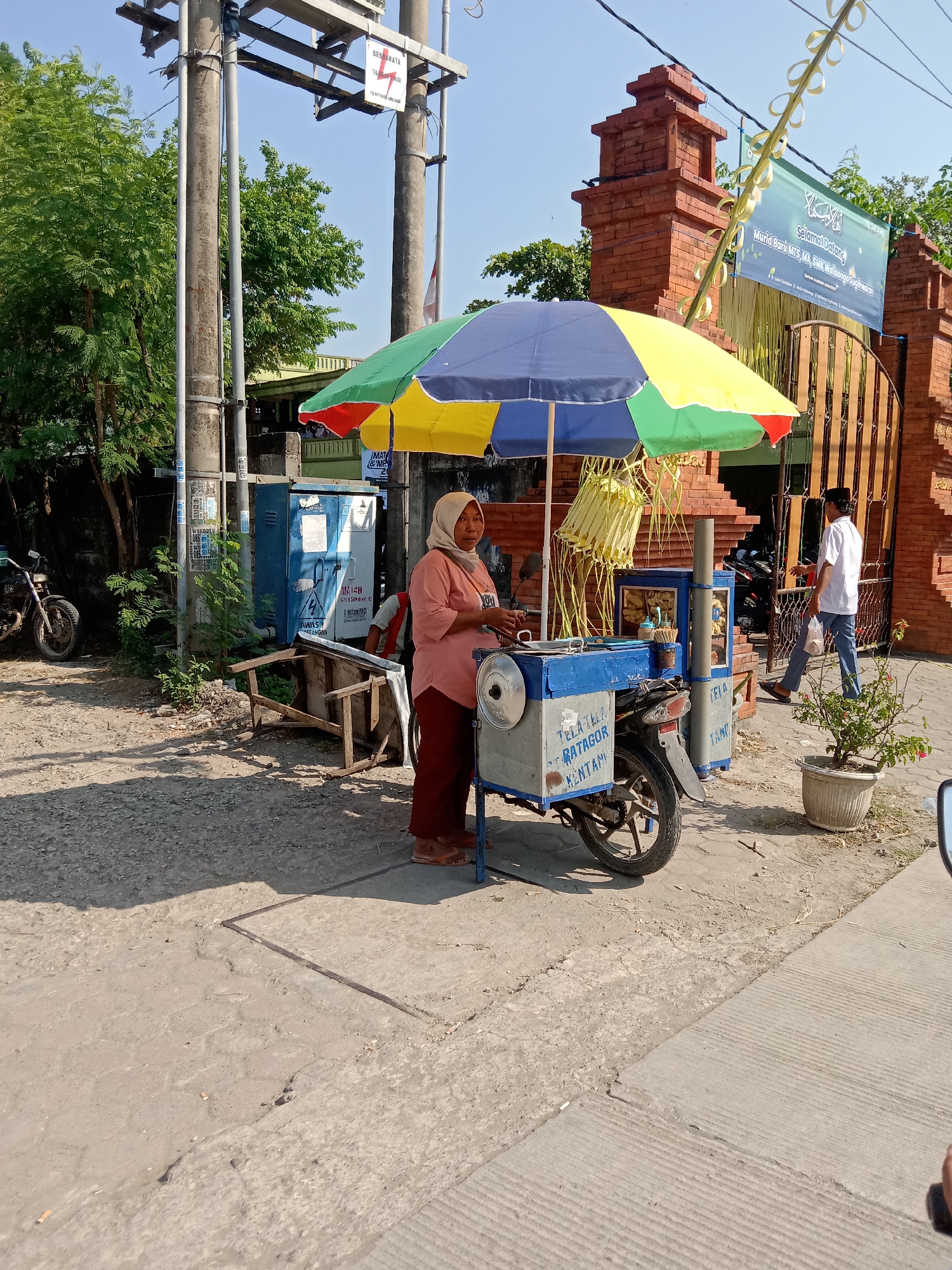 The image depicts an outdoor scene in the city of Sugihwaras, Indonesia. A woman wearing a hijab and a salmon-colored shirt stands next to a food cart under a multi-colored umbrella. The food cart is mounted on a motorcycle and appears to be selling snacks or drinks, with various items displayed in its compartments. The cart has text on its side, including "TELA TELA RATHOOR KENTANG". In the background, a brick gate with an ornate design is visible, adorned with a banner that reads "SELAMAT DATANG" and "Murid Baru MTs. SMK Walisongo Sugihwaras". A man in a white shirt and blue pants walks away from the gate, carrying a plastic bag. To the left of the food cart, a motorcycle is parked. The environment is sunny, with trees and power lines present. The ground is paved with concrete and gravel.