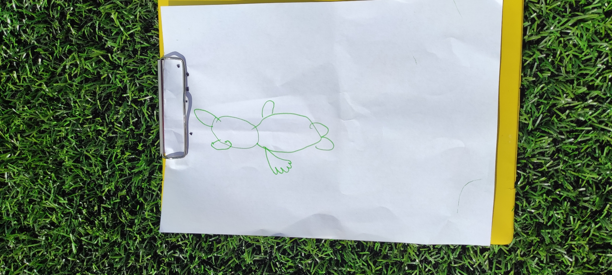 A sketch of a teddy bear in green marker on a white sheet of paper is held by a silver clip to a yellow clipboard. The clipboard rests on a green artificial turf surface, suggestive of a sports field or playground. The scene appears to be outdoors under daylight, with no distinct city landmarks visible. The location is not specified as Noviy, Russia.