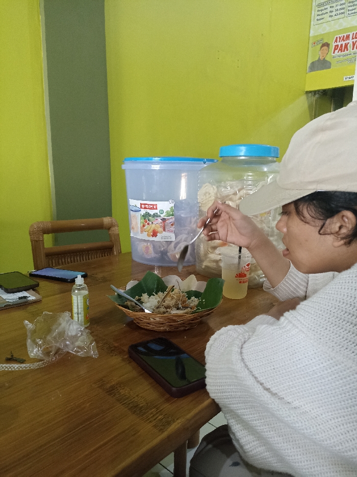 The image depicts a casual dining scene in an indoor setting, likely a local eatery or warung in Tulungagung, Indonesia. The central subject is a person, appearing to be a young woman, viewed from the right side, wearing a white knitted sweater and a light-colored baseball cap. She is actively eating a meal, holding a spoon near her mouth, focused on her food.
Her meal consists of rice and what appears to be shredded chicken or meat, served on banana leaves nested within a woven bamboo basket. A clear plastic cup with a light beverage and a straw is placed next to her food. On the wooden table, alongside the meal, are two smartphones, a small bottle of hand sanitizer, and a crumpled plastic bag. In the background, two large plastic containers are visible; one is clear and bears a "Lion Star" brand logo with a fruit graphic, while the other contains white, flat crackers or krupuk.
The walls are painted a vibrant lime green, with a darker green vertical accent strip. A bamboo-style chair is partially visible on the left. On the upper right wall, parts of a menu and signage are visible. The discernible text includes pricing: "Reguler Rp. 30.000", "Medium Rp. 38.000", and "Super Rp. 43.000". Below this, text reads "AYAM LE PAK Y" (likely referring to "Ayam Lele Pak Y", a common Indonesian dish/restaurant type) and "STAFF". The scene conveys a relaxed, everyday dining experience.