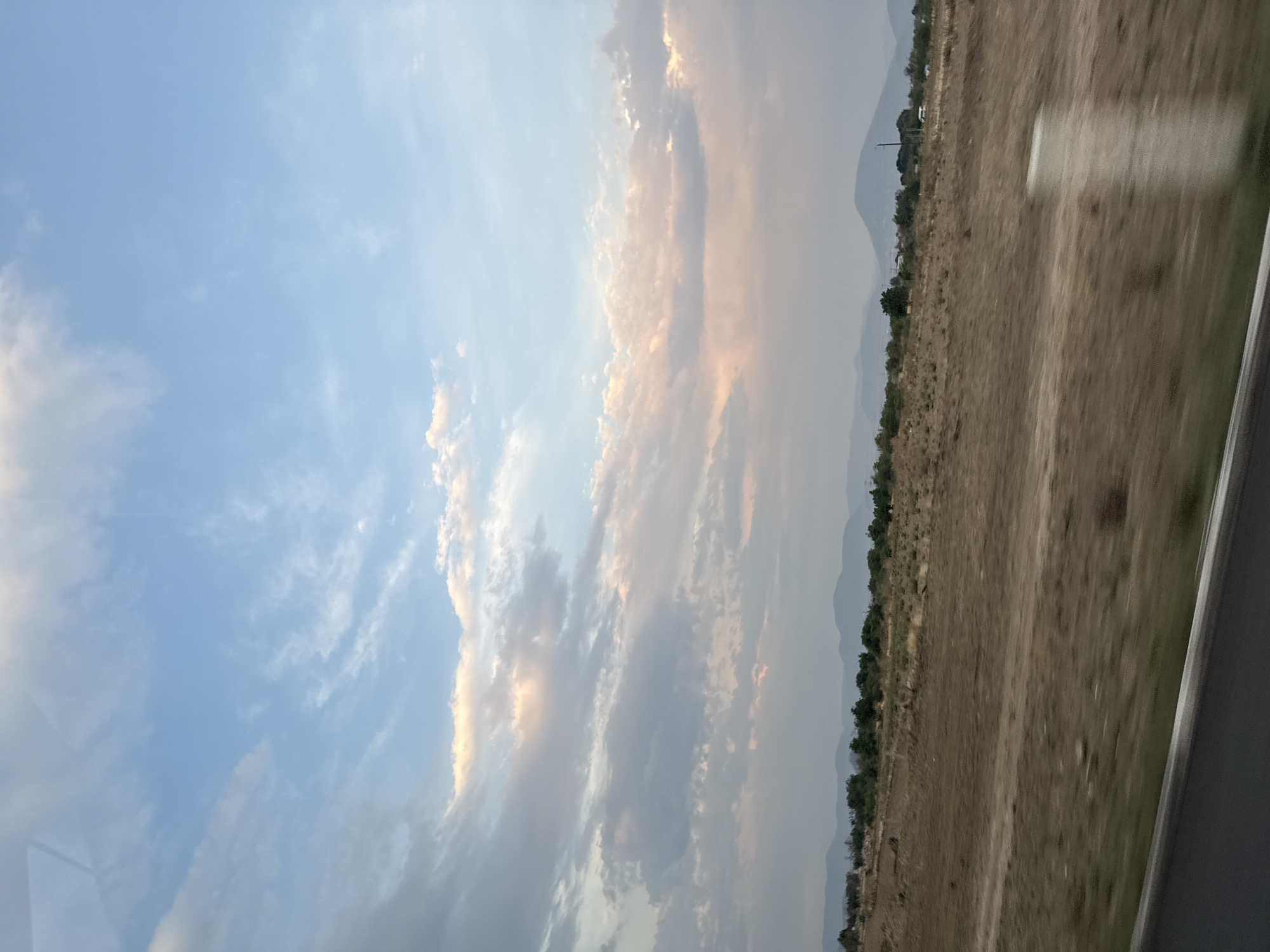 The image depicts a landscape with a blue sky containing wispy clouds and some golden tinges at the horizon. There is a mountain range in the distance. The foreground consists of dry, brown terrain interspersed with vegetation. A road or highway is visible at the bottom right corner. The scene is potentially located near Metepec, Mexico, based on geographic cues.