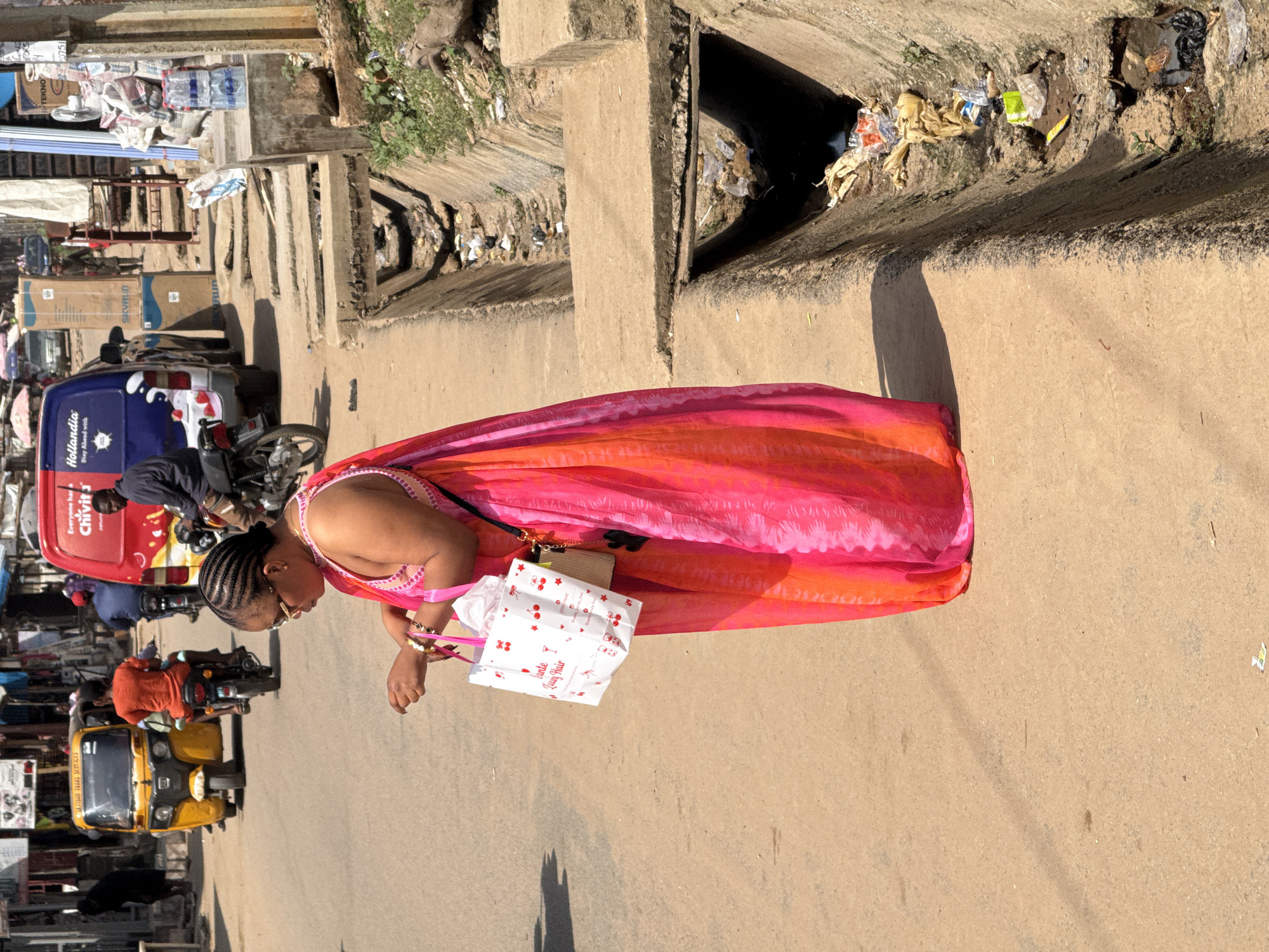 A woman in a bright pink and orange patterned dress, holding a white shopping bag adorned with cherries, walks along a dusty road in Abuja, Nigeria. The woman is wearing gold-rimmed glasses and has her hair braided in cornrows. In the background, a yellow auto-rickshaw is parked on the side of the road, and several people are visible in the distance, some on motorcycles and others walking. A red and blue commercial van with "Hollandia" branding is also present. The street appears to be a busy urban environment with shops and market stalls lining one side of the road, where various goods are displayed. A prominent concrete drainage channel filled with debris runs along the edge of the road. The overall scene depicts a vibrant streetscape in a Nigerian city, characterized by the presence of vehicles, pedestrians, and commercial activity.