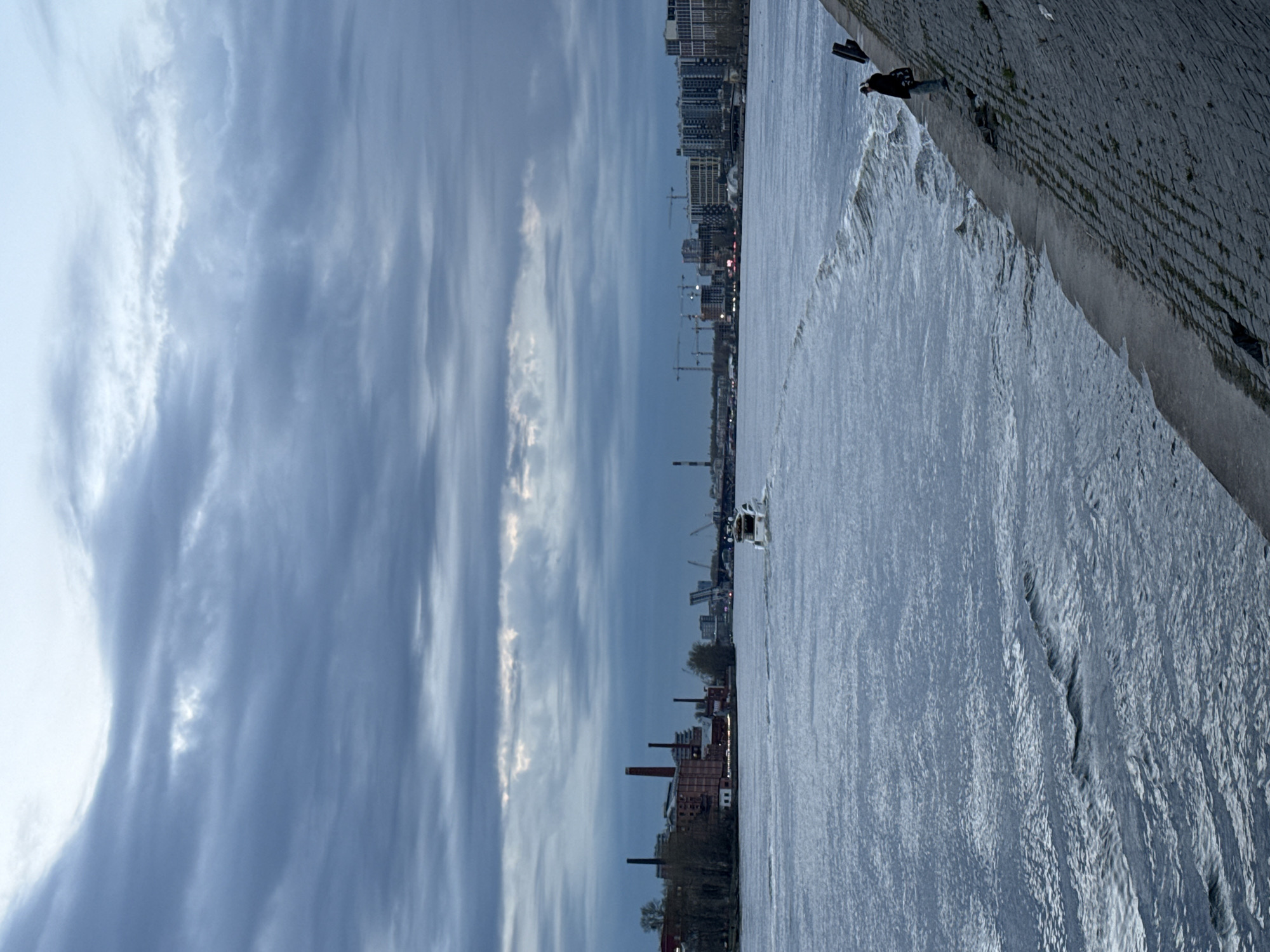 This image, set in Saint Petersburg, Russia, captures a wide river, likely partially frozen or filled with slush, under a cloudy sky. The scene is oriented to show an expansive body of water in the foreground and mid-ground, with an urban cityscape stretching along the far bank and a stone embankment on the near bank.
The water surface is textured with patterns of slush or thin ice, suggesting cold weather, possibly late winter or early spring. On the far bank, a dense skyline features a mix of multi-story residential or commercial buildings, alongside older industrial structures marked by tall red-brick chimneys. Several construction cranes dot the skyline, indicating ongoing development in the area.
Along the near bank, a robust, textured stone or brick embankment is visible. Two individuals are present on this embankment towards the right side of the frame; one appears to be sitting or leaning, while the other stands beside them. They seem to be observing the river or the city across it.
The sky is largely covered with long, wispy, and streaky clouds, contributing to an overcast atmosphere. The lighting is diffused and soft, typical of a cloudy daytime. The overall mood is calm and somewhat muted, reflecting the cool weather and urban environment. No discernible text is visible in the image.