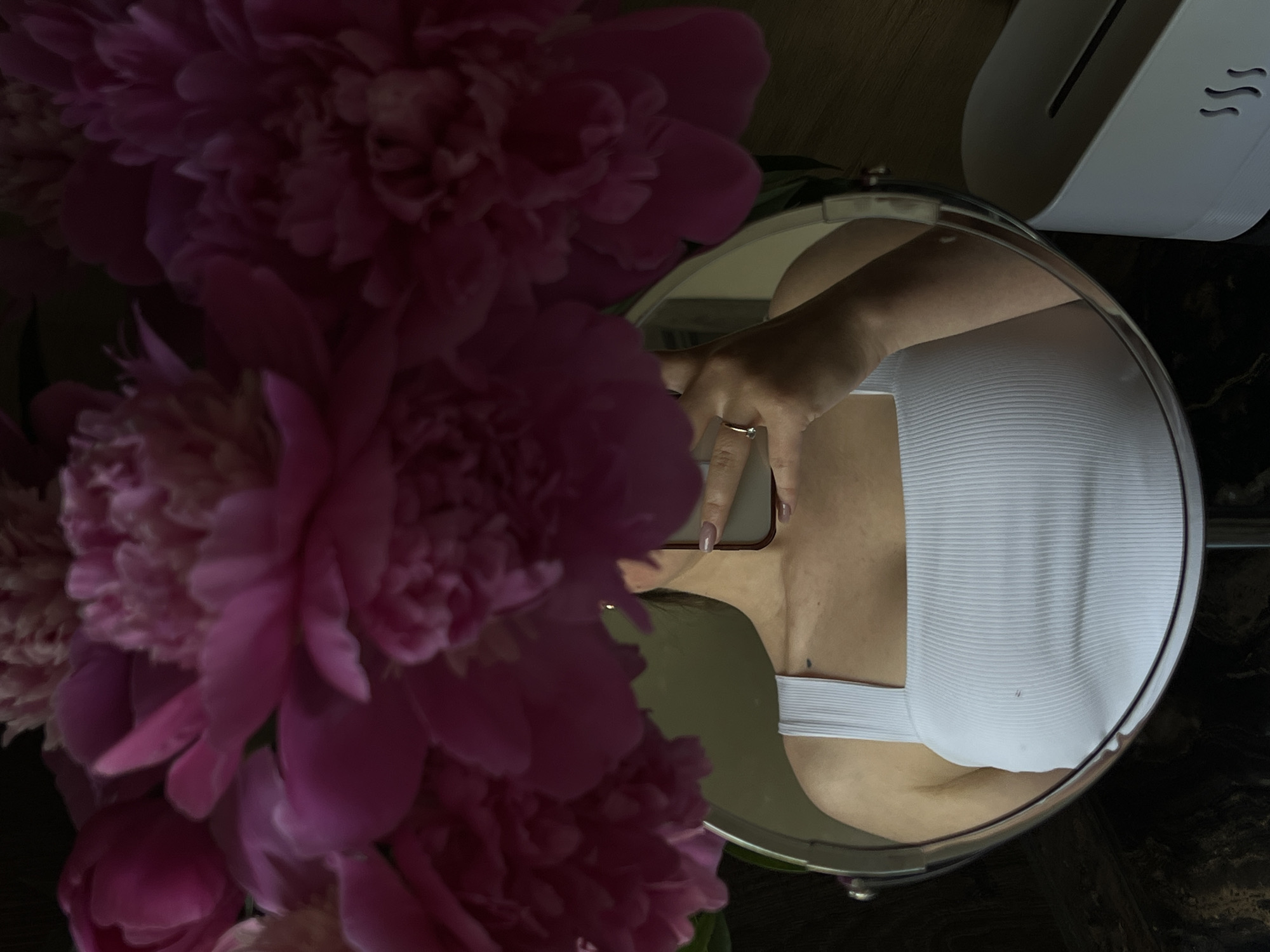 A close-up, low-angle shot shows a bouquet of pink peonies positioned above a round mirror that reflects a person's upper body. The person is wearing a white ribbed tank top with a square neckline and is holding a smartphone in their right hand, capturing the reflection. A ring is visible on the ring finger of the right hand. The mirror is on a dark, reflective surface, possibly a table. To the right of the mirror, a white tissue box with a slot for dispensing tissues is visible. The background is dark and out of focus, with the peonies dominating the upper portion of the frame. There is no information in the image to suggest a location in Ternopil, Ukraine.