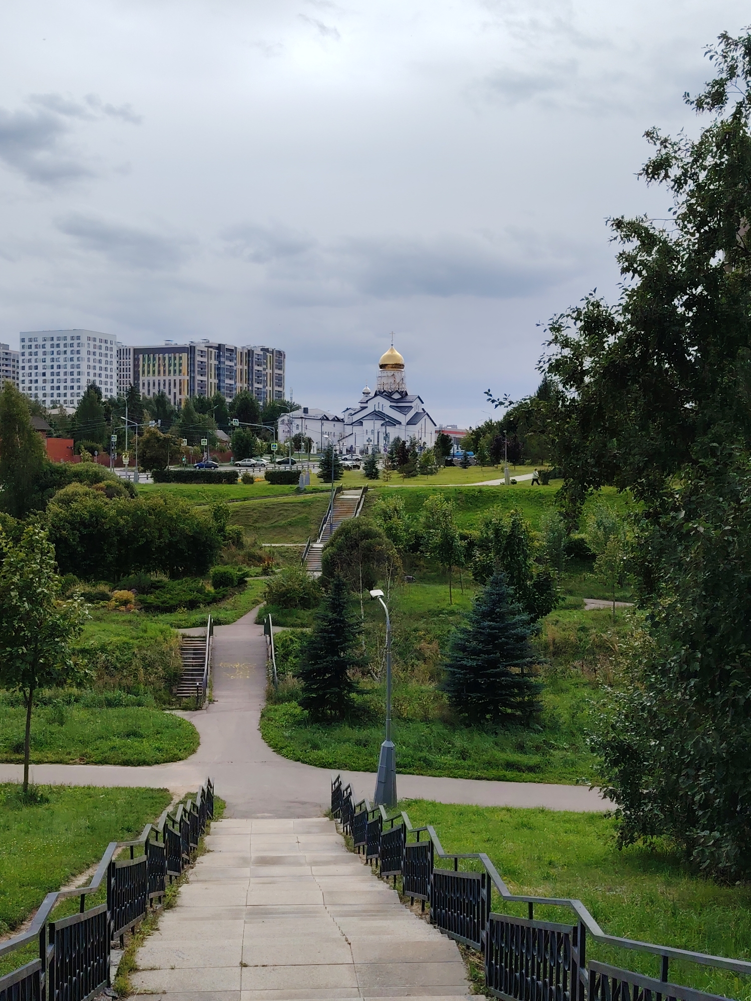 The image shows a park scene in Zelenograd, Russia. In the foreground, a set of concrete stairs with black metal railings leads down. The path continues with a paved walkway curving to the left and a set of stairs ascending further into the park. The park is characterized by lush green grass, trees, and some smaller evergreen trees. In the middle ground, a pedestrian walks along a curved path. In the background, a white Orthodox church with a golden dome is visible. To the left of the church are modern, multi-story residential buildings with colorful facades. The sky is overcast with gray clouds.
