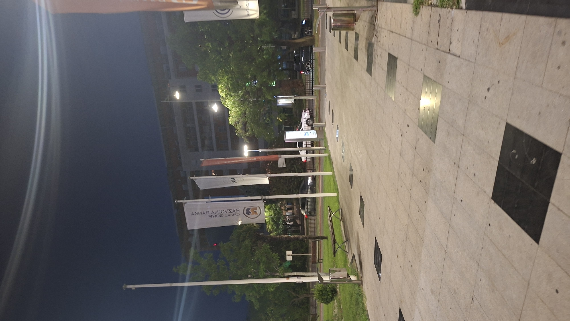 The image depicts an outdoor scene at night in Podgorica, Montenegro. The foreground features a paved walkway with square, dark inlays. To the left, there is a grassy area with a bench and a trash receptacle. Several flags are visible, some with text that appears to be in Cyrillic script, mounted on poles. A white vehicle and a dark-colored vehicle are driving on a road in the mid-ground, separated from the walkway by a fence. In the background, a multi-story building with illuminated windows is visible, along with trees and streetlights. The sky is dark, suggesting it is nighttime, and there is some lens flare visible in the upper portion of the image.