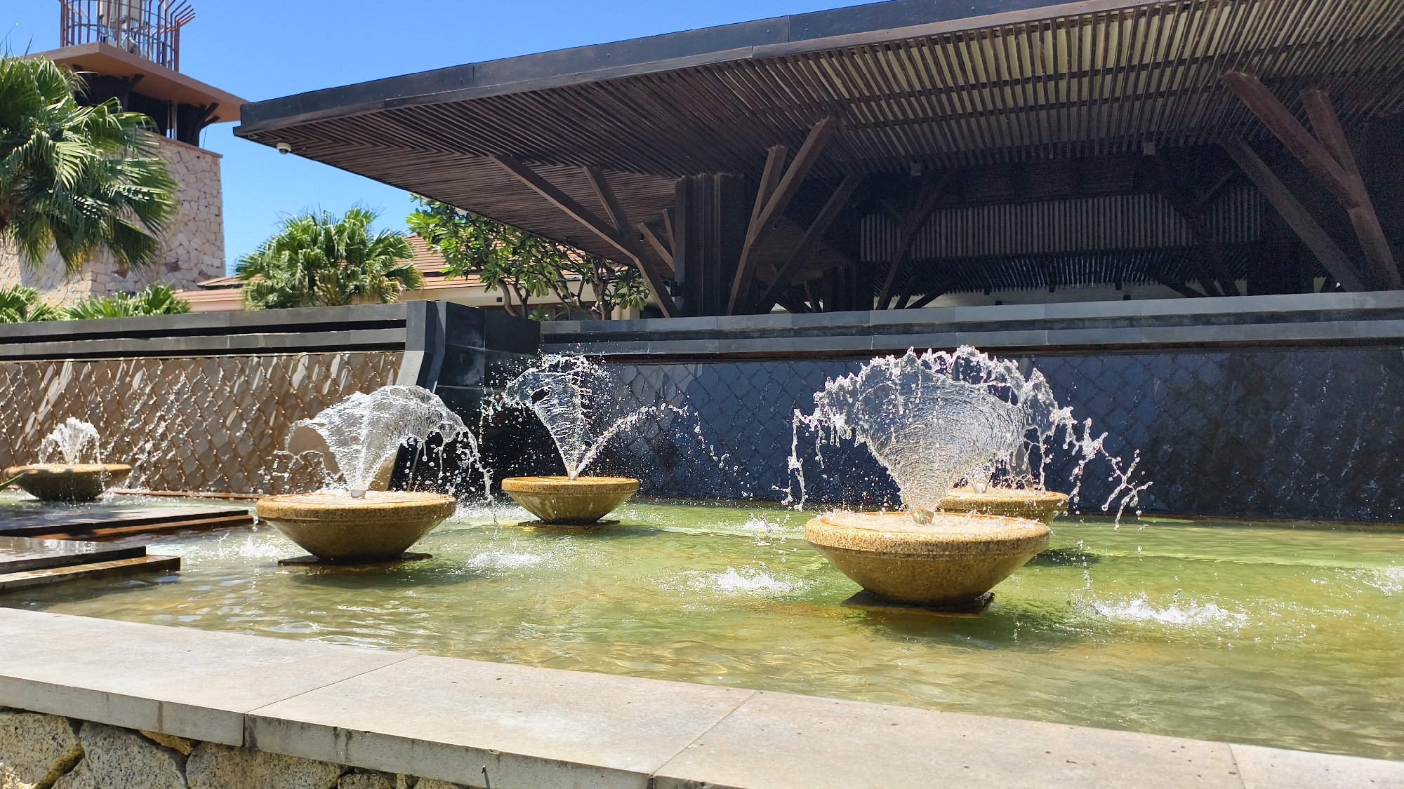 The image depicts an outdoor water feature with multiple tiered fountains in a pool of greenish water. The fountains are made of a light-colored, textured material and are spraying water into the air, creating splashes and droplets. Behind the fountains is a dark, textured wall, possibly made of stone or tile, with a lighter-colored horizontal band above it. Overhanging the scene is a large, dark wooden roof structure supported by angled beams. In the background, there are palm trees and a stone tower, suggesting a tropical or resort-like environment. The sky is a clear blue. The scene appears to be located in Nha Trang Ward, Vietnam.